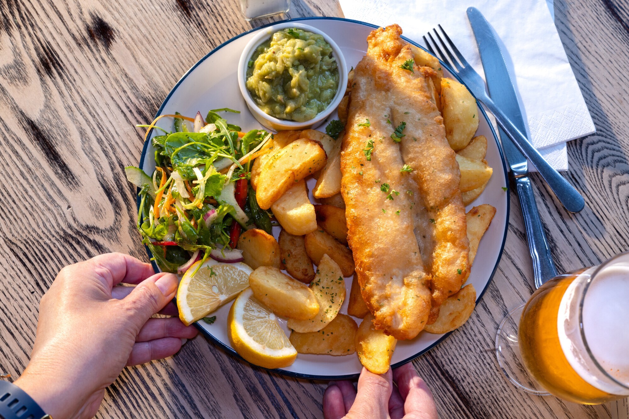 Eine Hand greift nach einem Teller Fish and Chips mit Beilagen auf einem Holztisch bei Sonnenschein.