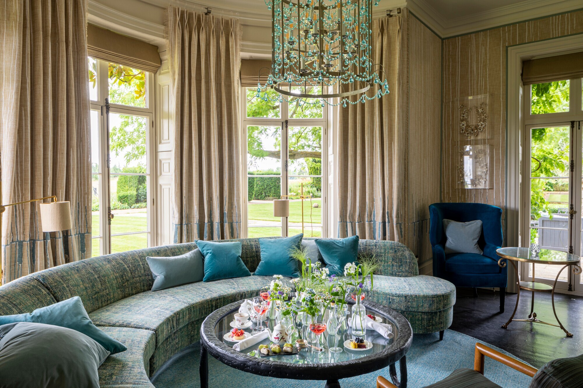 Luxuriöser Salon mit Kronleuchter und geschwungenem Sofa vor Panoramafenstern mit Blick in einen Garten. Luxuriöser Salon mit Kronleuchter und geschwungenem Sofa vor Panoramafenstern mit Blick in einen Garten.