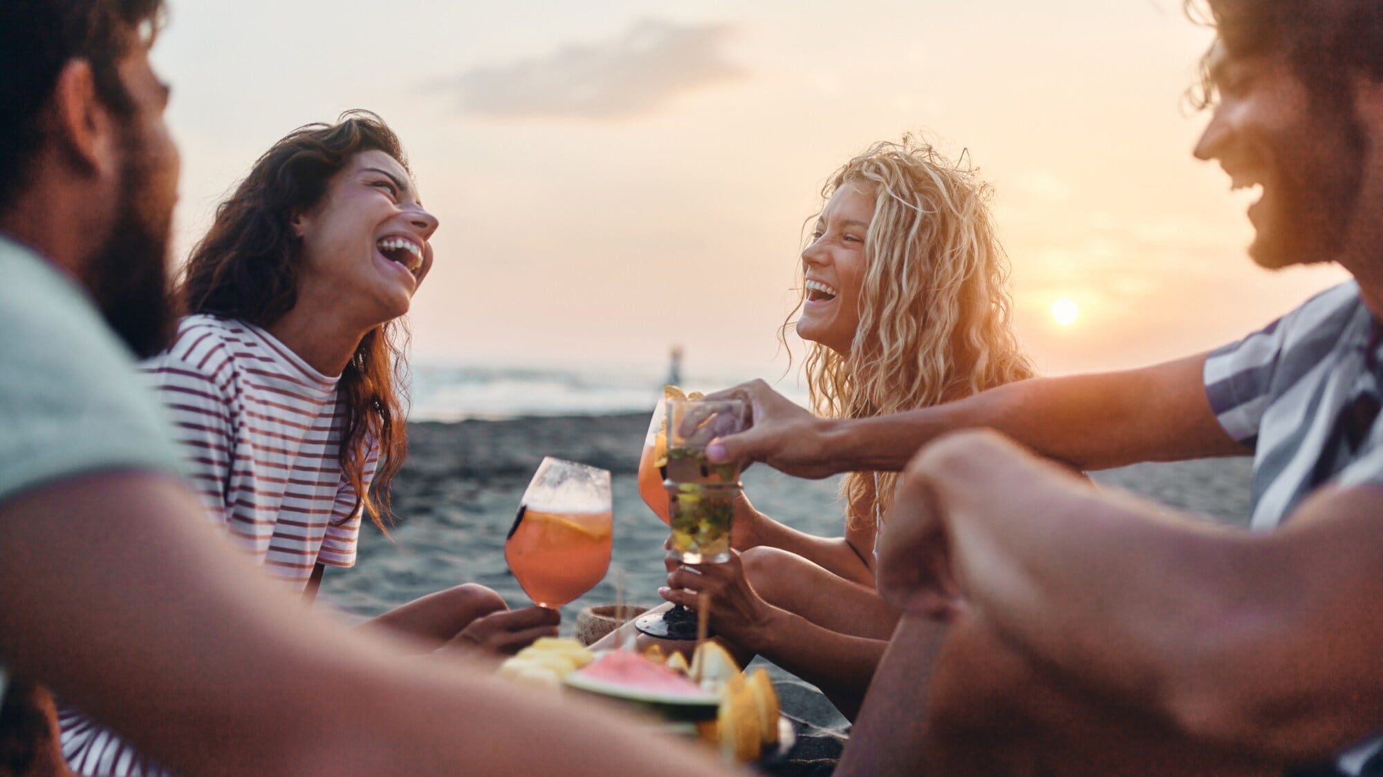 Eine Gruppe von vier fröhlichen Personen sitzt am Meer bei Sonnenuntergang mit Drinks und Snacks. Eine Gruppe von vier fröhlichen Personen sitzt am Meer bei Sonnenuntergang mit Drinks und Snacks.