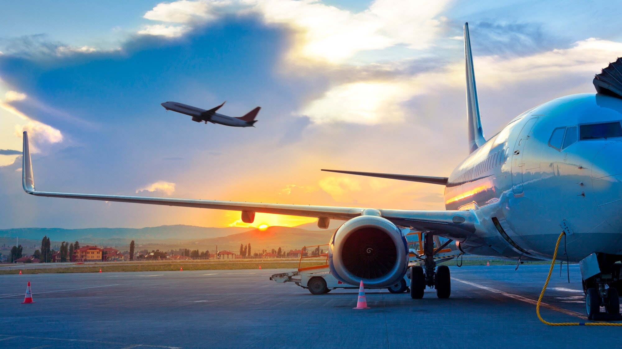 Ein Flugzeug am Himmel über einem geparkten Flugzeug am Flughafen vor einer hügeligen Landschaft bei Sonnenuntergang. Ein Flugzeug am Himmel über einem geparkten Flugzeug am Flughafen vor einer hügeligen Landschaft bei Sonnenuntergang.