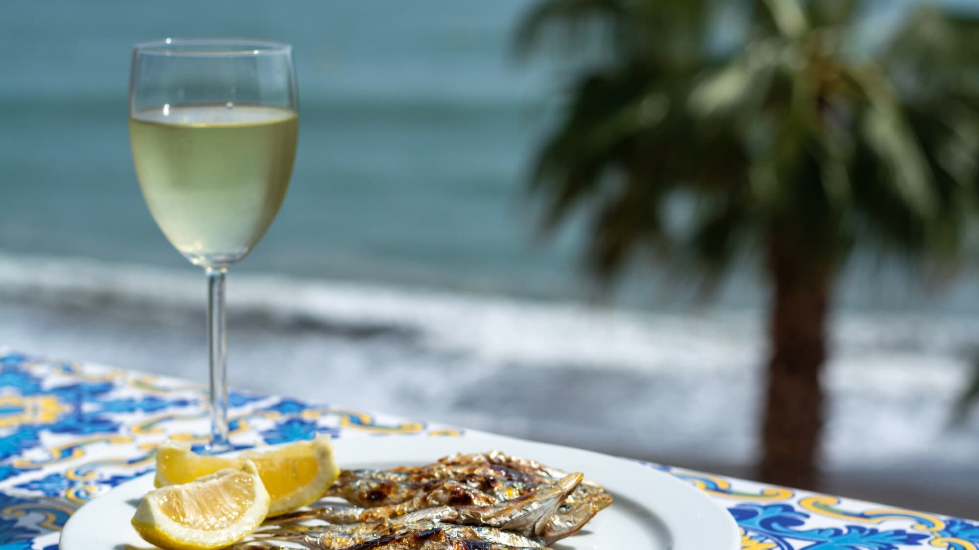 Gegrillte Sardinen auf einem weißen Teller neben einem Glas Weißwein, dahinter Meer mit Palme in Unschärfe. Gegrillte Sardinen auf einem weißen Teller neben einem Glas Weißwein, dahinter Meer mit Palme in Unschärfe.