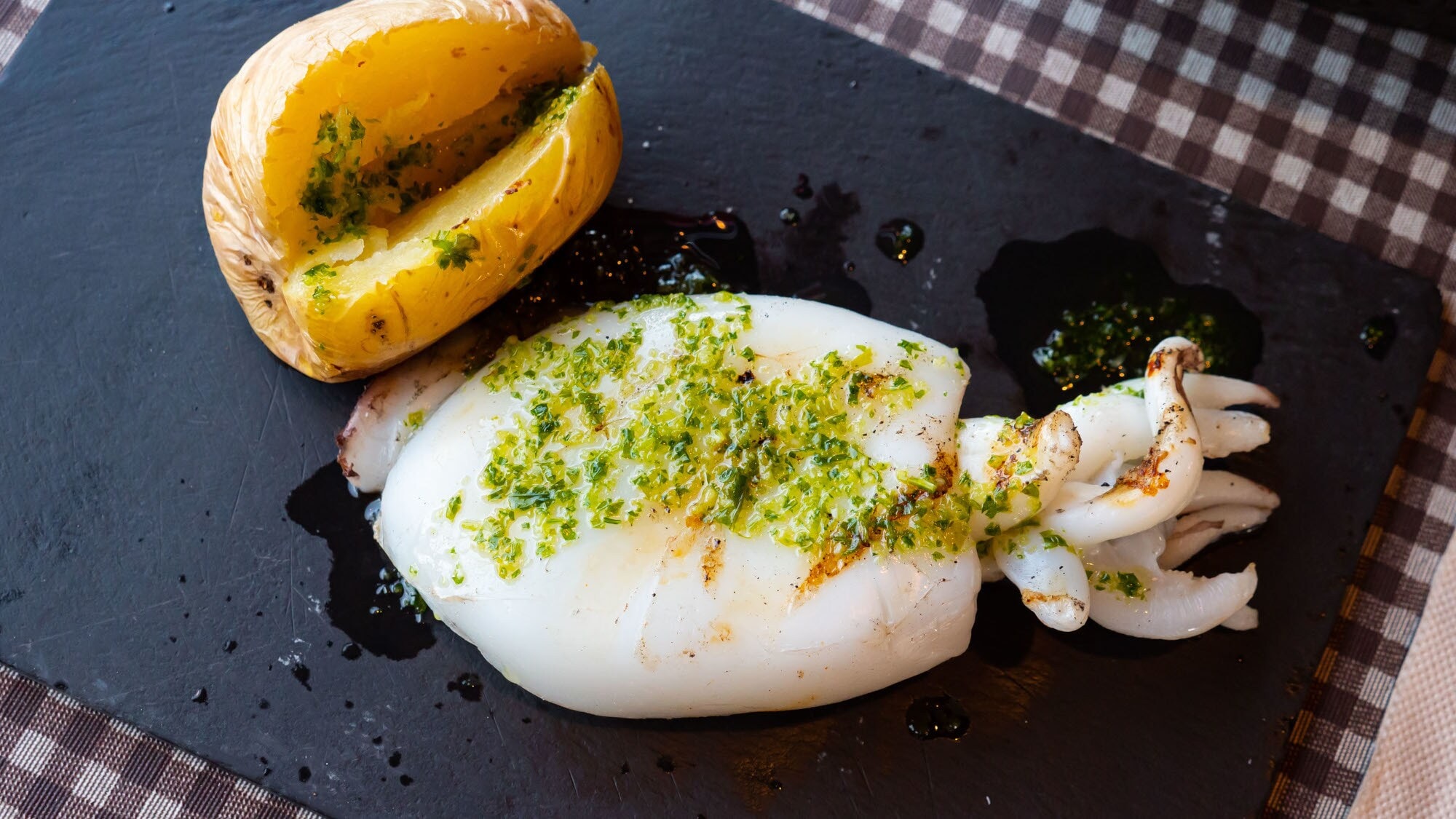 Aufsicht eines zubereiteten Tintenfisches auf einem schwarzen Küchenbrett, daneben eine gebackene Kartoffel. Aufsicht eines zubereiteten Tintenfisches auf einem schwarzen Küchenbrett, daneben eine gebackene Kartoffel.