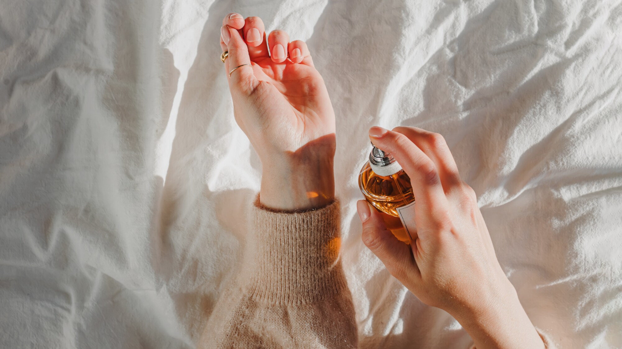 Nahaufnahme der Hände einer Person über einem weißen Bettlaken; in einer Hand hält sie eine orange-braune Parfumflasche, mit der sie das andere Handgelenk besprüht.