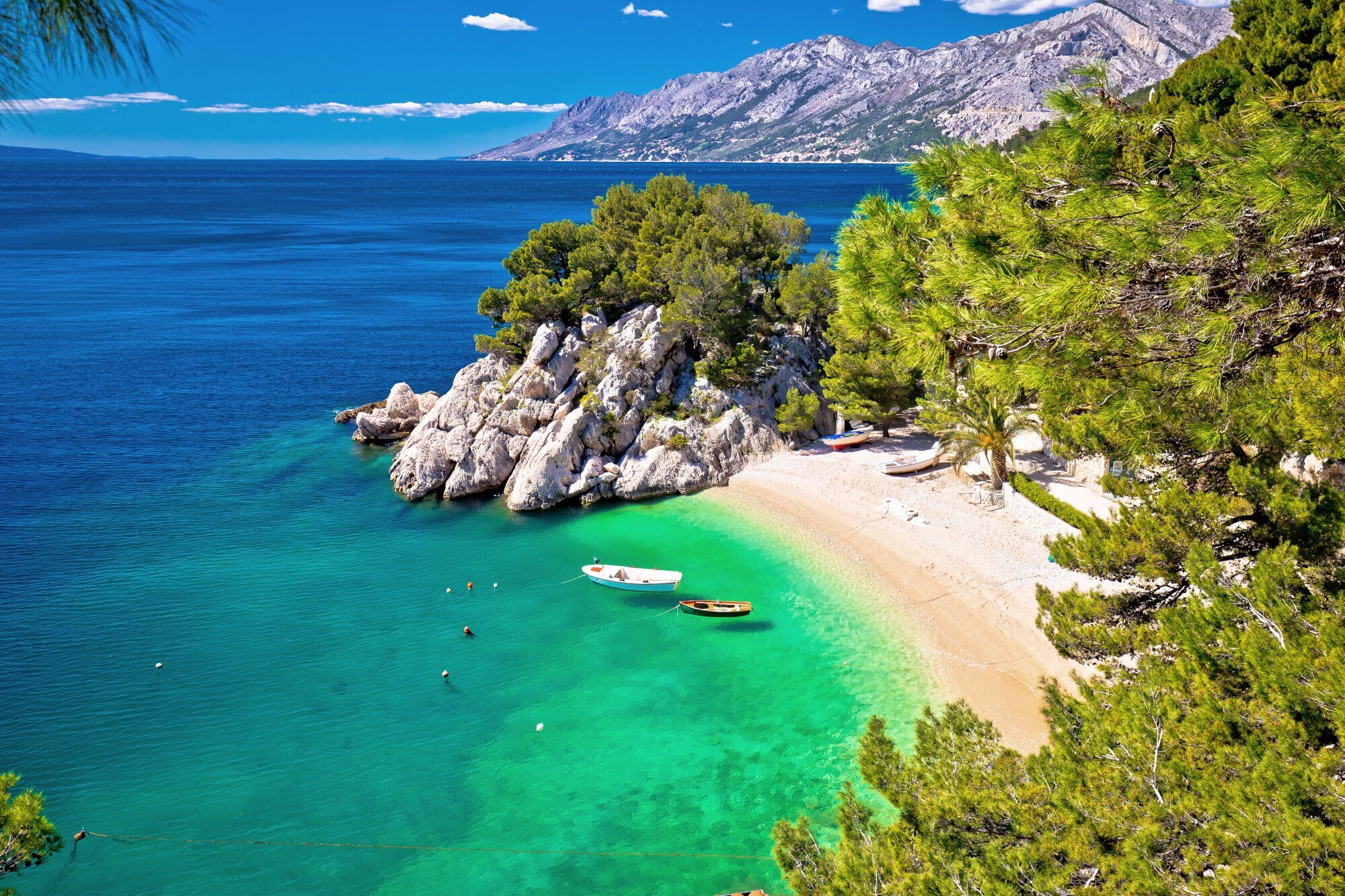 Kleine Sandbucht mit Fischerbooten im türkisblauen Meer, umgeben von Felsen und Kiefernwald.