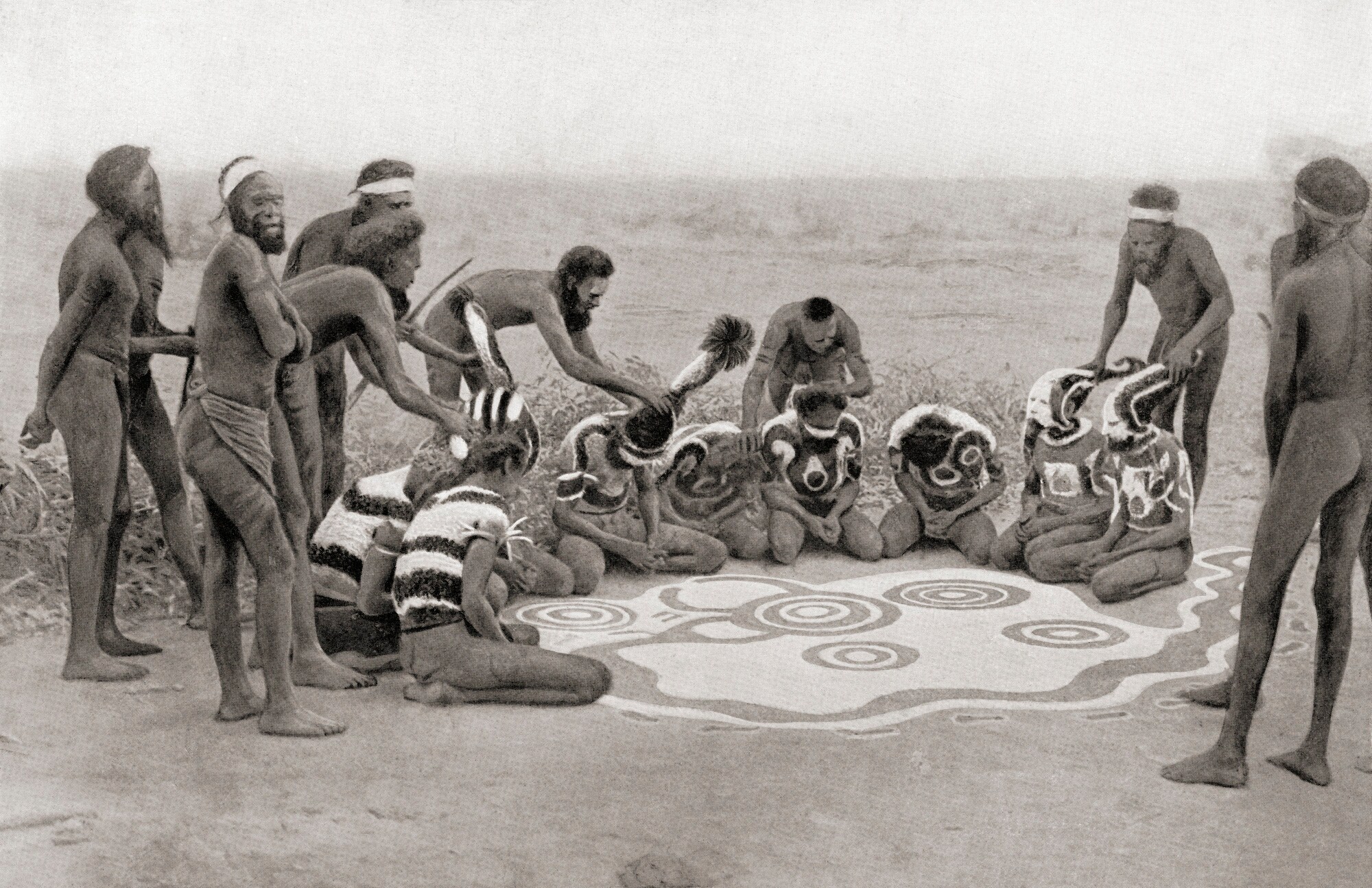 Eine Gruppe Aboriginals mit Körper- und Bodenbemalung während einer Zeremonie, Schwarzweißaufnahme.