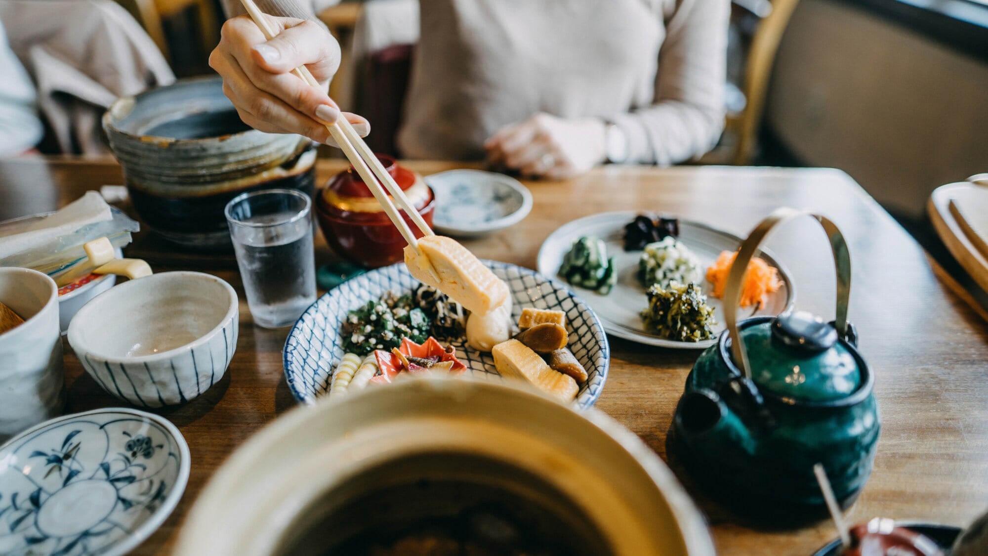 Auf einem Tisch stehen mehrere Schälchen mit verschiedenen asiatischen Gerichten. Eine unkenntliche Person hält mit Stäbchen etwas zu essen hoch. Auf einem Tisch stehen mehrere Schälchen mit verschiedenen asiatischen Gerichten. Eine unkenntliche Person hält mit Stäbchen etwas zu essen hoch.