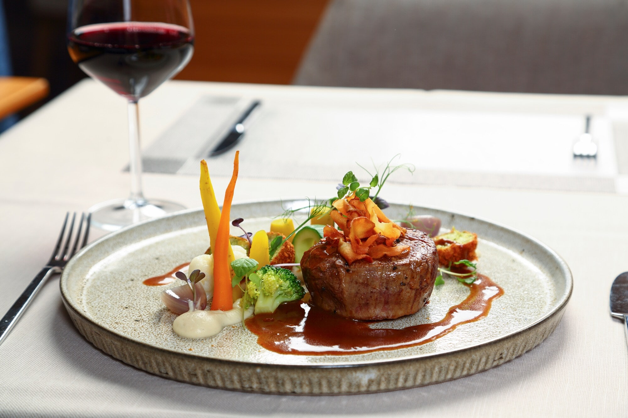 Ein Stück Steak an Gemüse, angerichtet auf einem Teller neben einem Glas Rotwein auf einem weiß gedeckten Tisch. Ein Stück Steak an Gemüse, angerichtet auf einem Teller neben einem Glas Rotwein auf einem weiß gedeckten Tisch.