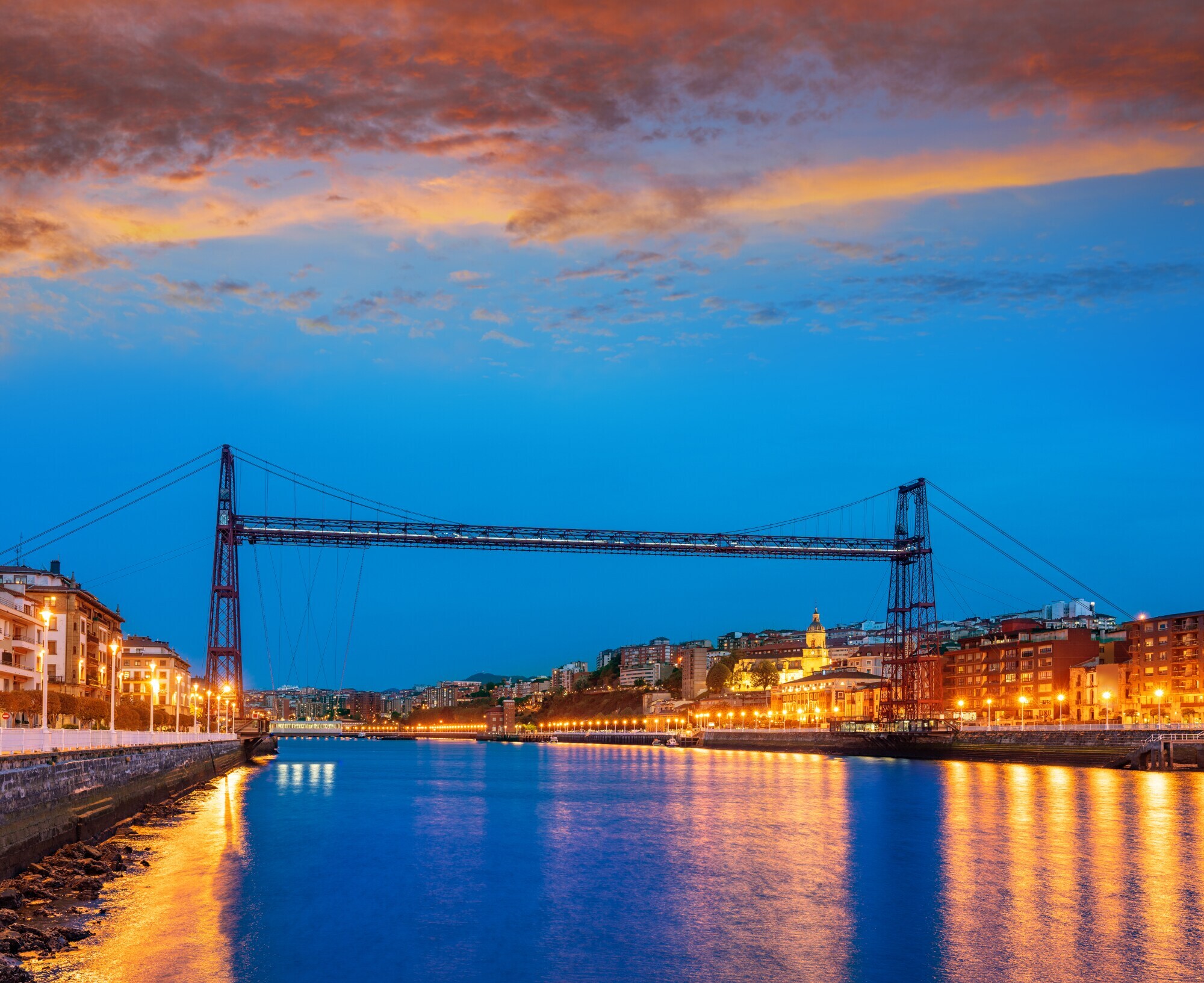 Die Puente de Vizcaya ragt bei Nacht über den Fluss, rechts und links befinden sich beleuchtete Häuser.