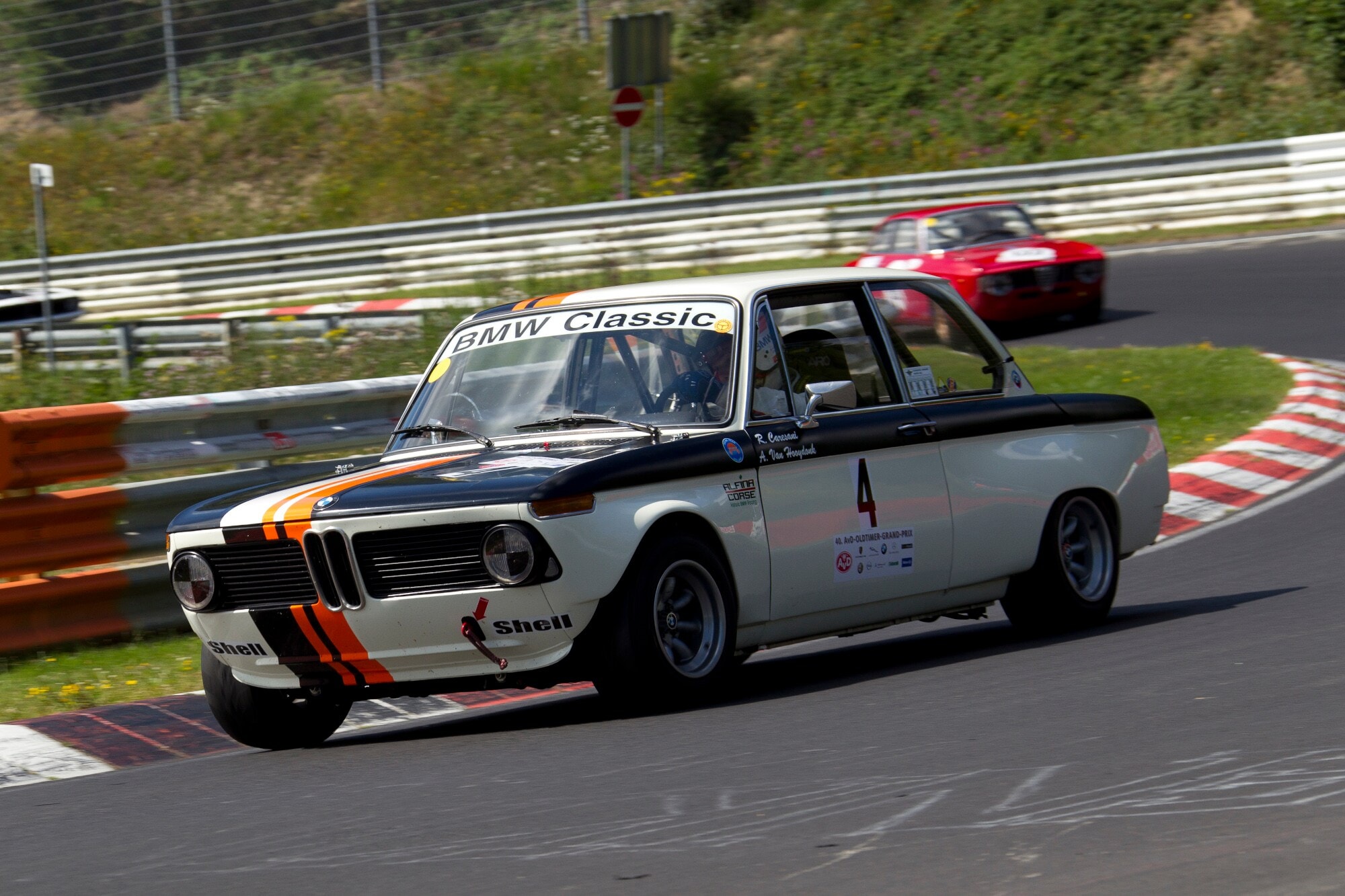Ein fahrender BMW 2002 ti Rennwagen in der Kurve einer Rennstrecke, dahinter ein weiterer Rennwagen. Ein fahrender BMW 2002 ti Rennwagen in der Kurve einer Rennstrecke, dahinter ein weiterer Rennwagen.