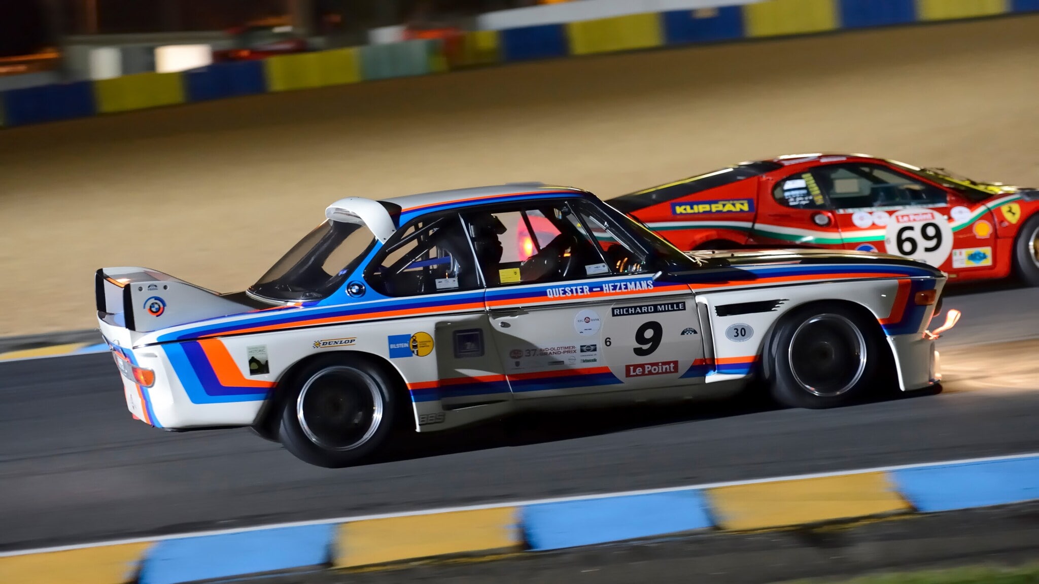 Ein fahrender BMW 3.0 CSL Rennwagen mit Rallyestreifen auf einer Rennstrecke vor einem anderen Rennwagen. Ein fahrender BMW 3.0 CSL Rennwagen mit Rallyestreifen auf einer Rennstrecke vor einem anderen Rennwagen.