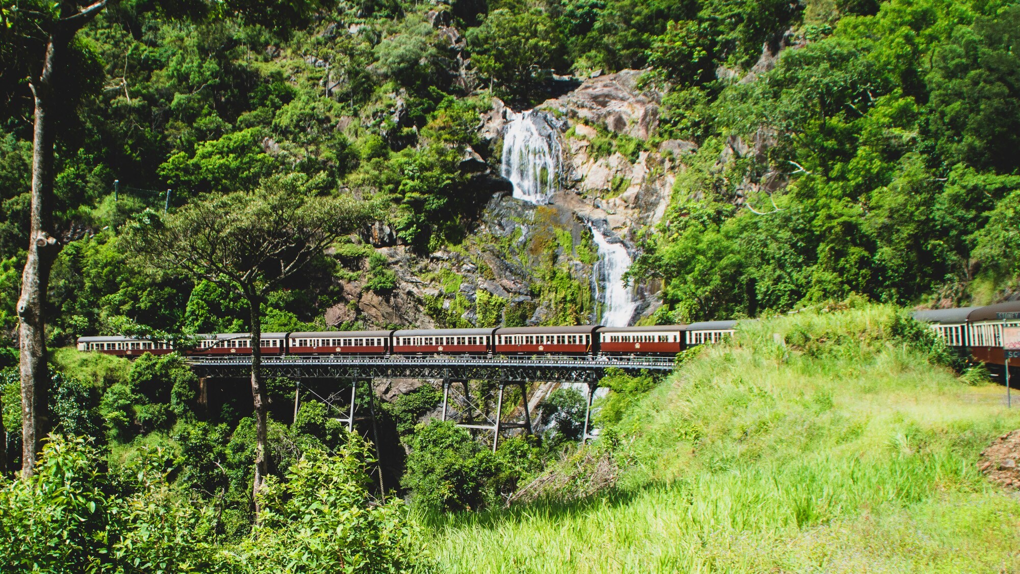 Eine historische Eisenbahn fährt über eine Brücke vor einem Wasserfall im Regenwald.