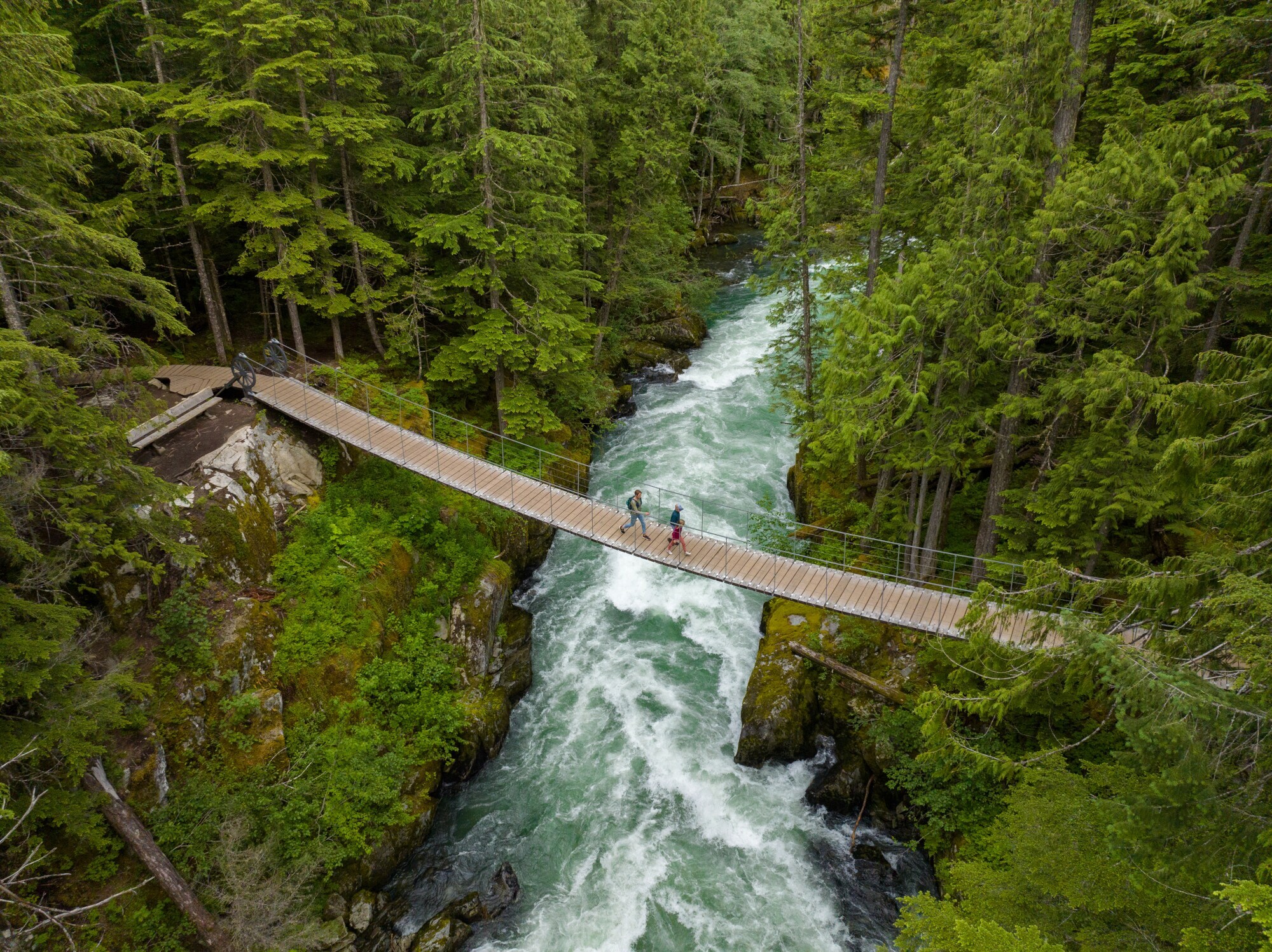 Luftaufnahme zweier Personen, die auf einer Hängebrücke einen wilden Fluss in einem Waldgebiet überqueren.