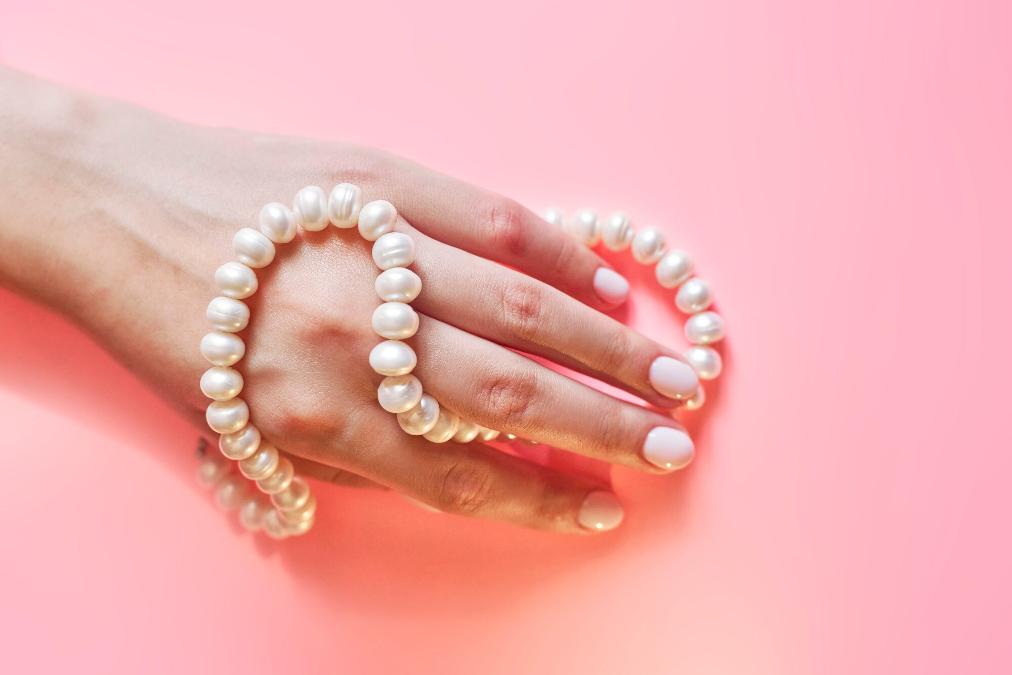 Eine Frauenhand mit Perlenkette umwickelt auf rosa Untergrund