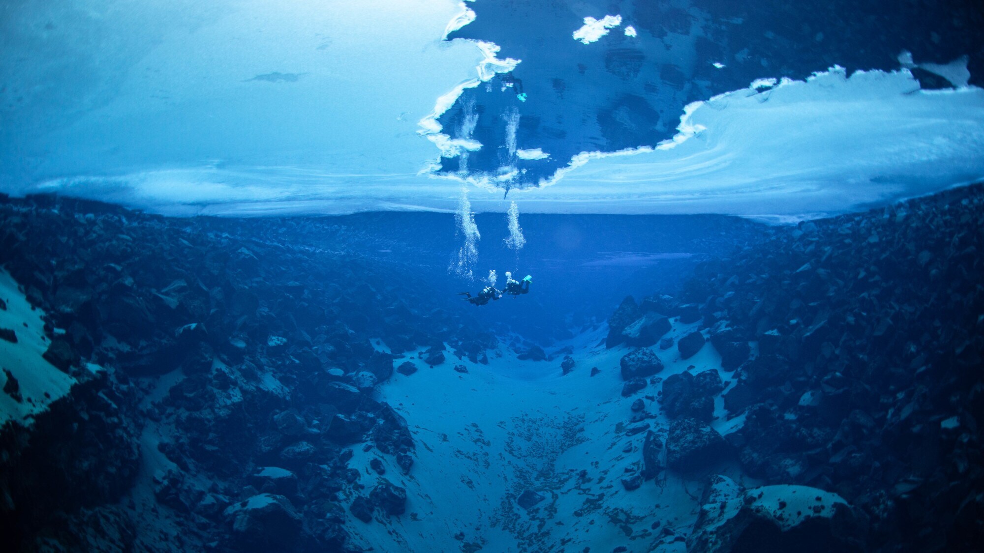 Zwei Personen in Tauchausrüstung tauchen durch ein Loch in der Eisdecke in einen zugefrorenen See.