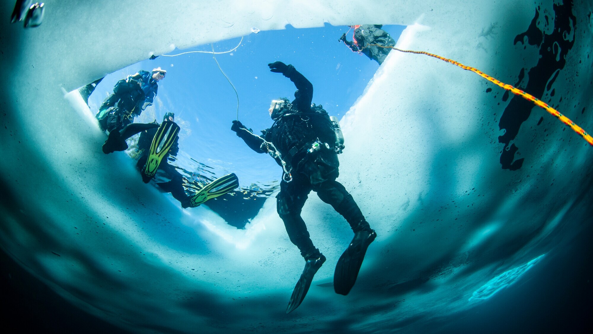 Eine Person in Taucherausrüstung, die aus dem zugefrorenen Wasser aus einem Loch in der Eisdecke auftaucht. Am Rand sitzt eine weitere Person.