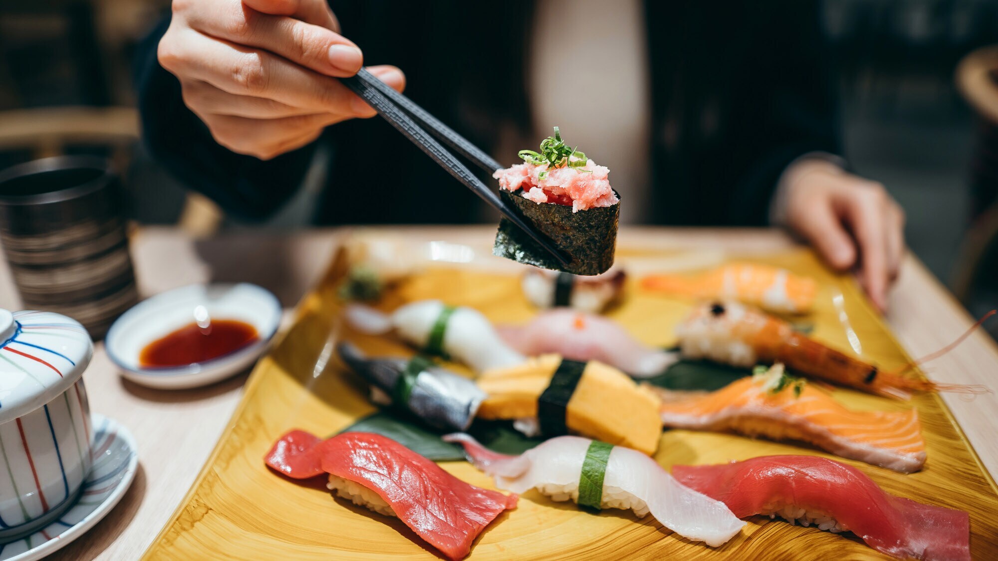 Eine Person hält eine Rolle Maki Sushi zwischen zwei Essstäbchen über einer Bambusplatte mit Nigiri auf einem Tisch in einem Restaurant.