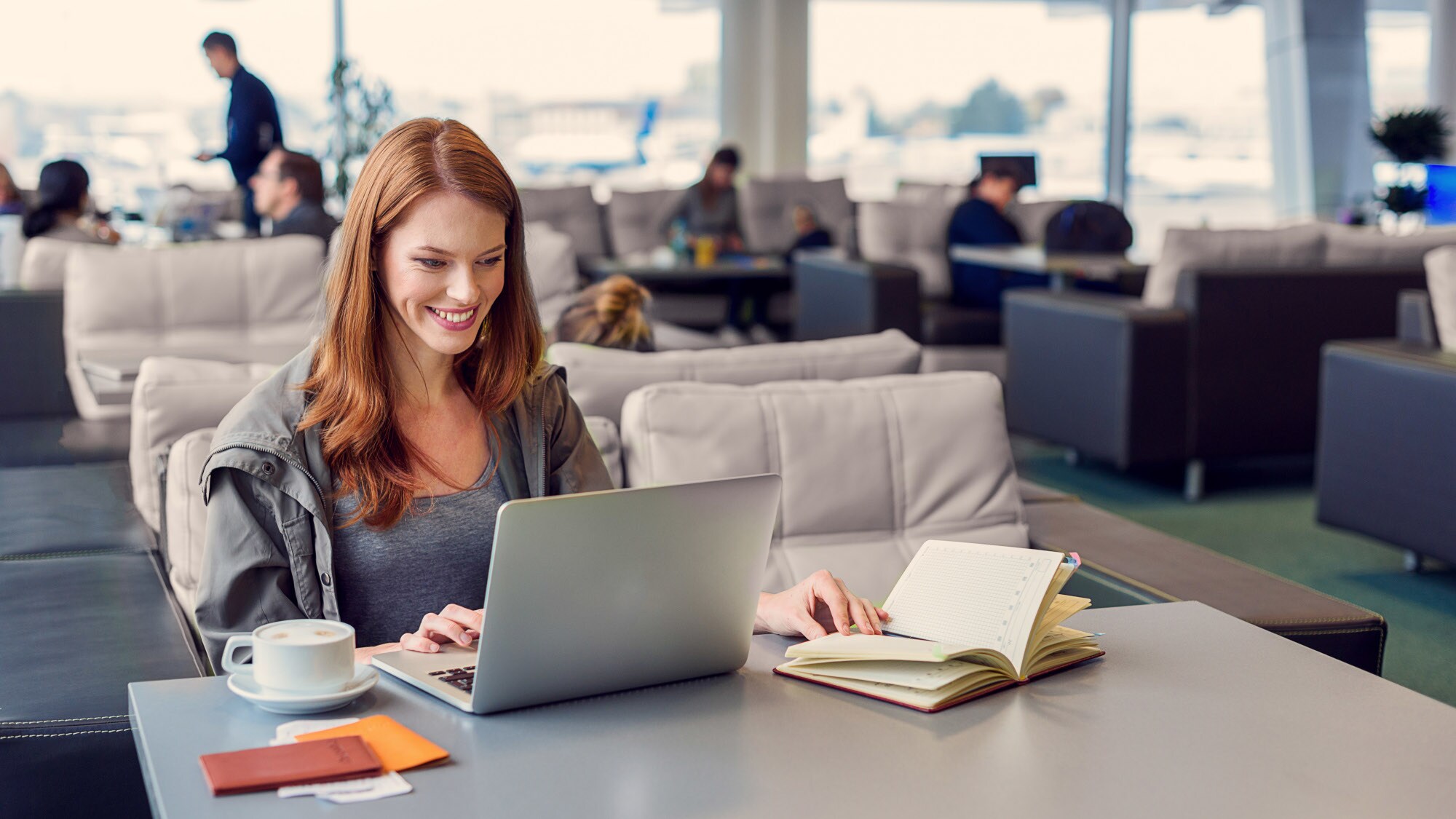 Eine lächelnde Frau arbeitet an ihrem Laptop an einem Tisch in einer Flughafen-Lounge. Eine lächelnde Frau arbeitet an ihrem Laptop an einem Tisch in einer Flughafen-Lounge.
