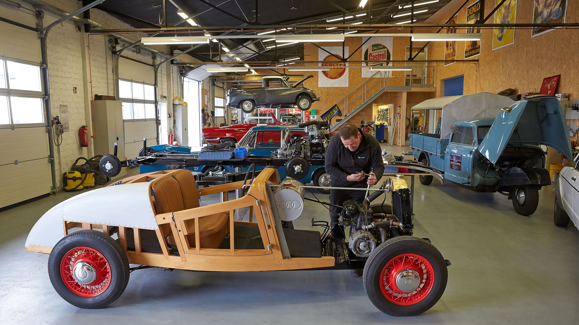 Ein Mechaniker arbeitet an einem Oldtimer mit Holzgestell in einer Werkstatt mit Ausstellungsraum. Ein Mechaniker arbeitet an einem Oldtimer mit Holzgestell in einer Werkstatt mit Ausstellungsraum.