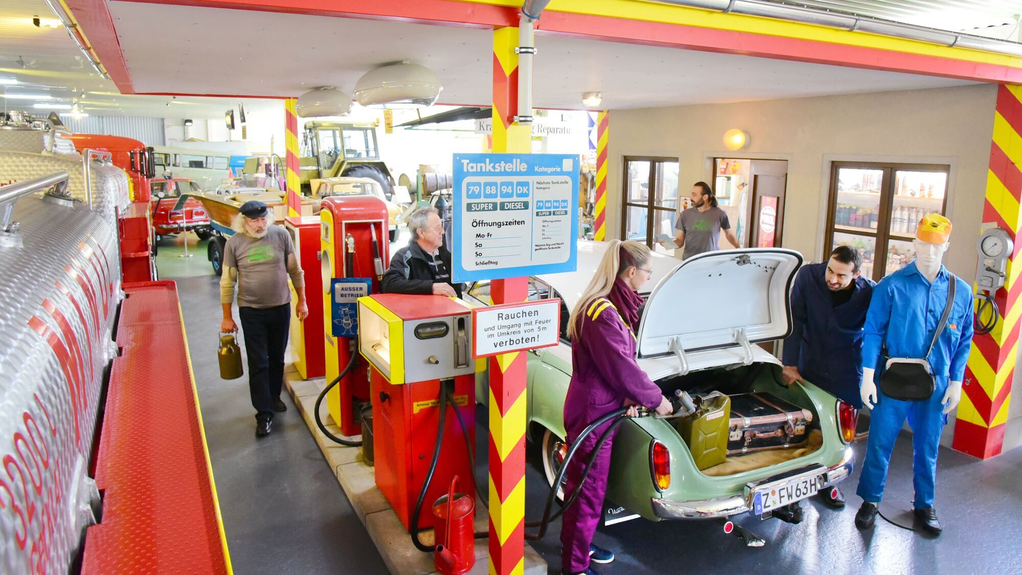Personen an einer nachgebildeten Tankstelle mit Oldtimern in einem Ausstellungsraum.