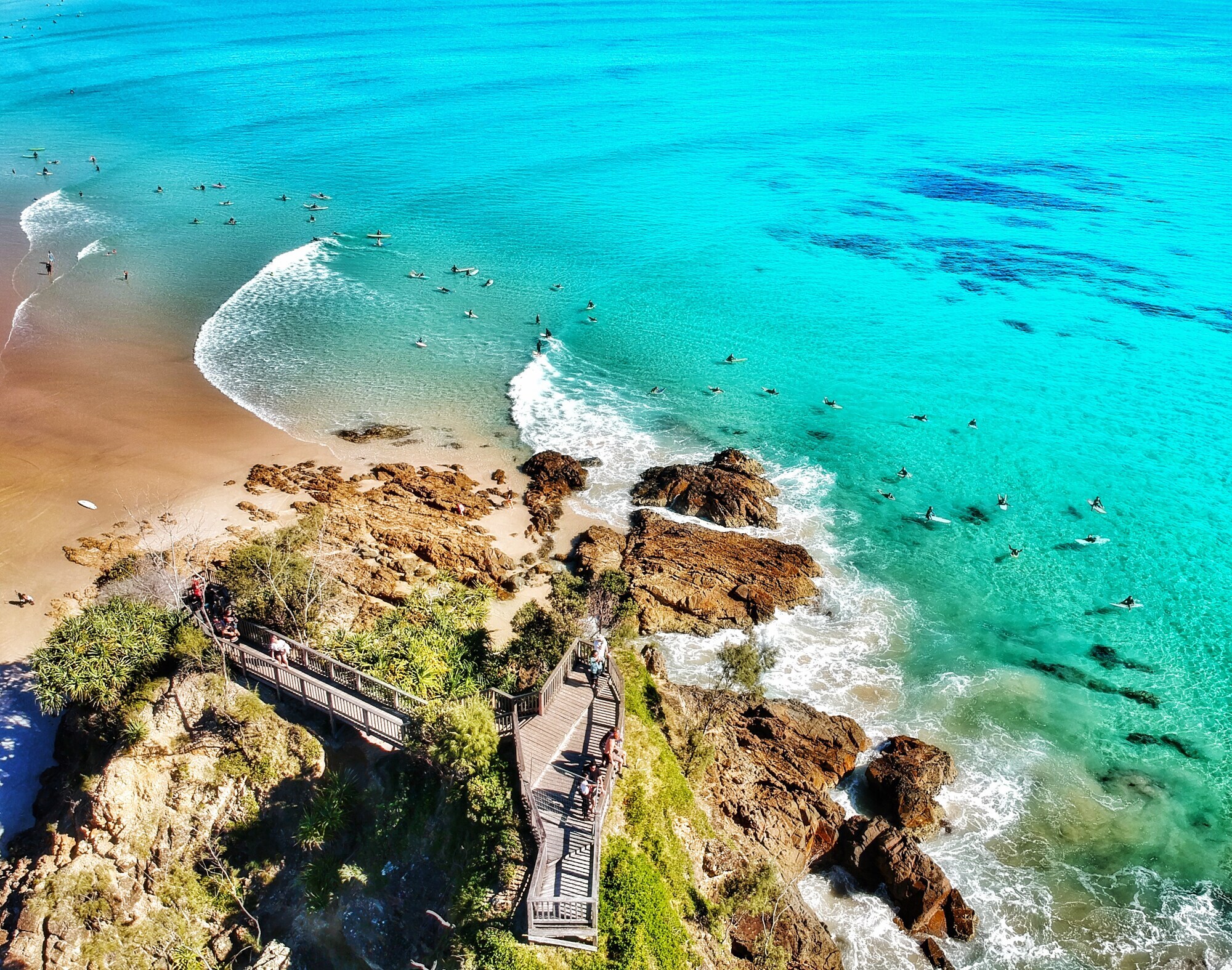 Aufsicht eines Küstenabschnittes mit Felsen und Sandstrand, dahinter zahlreiche Surfende im türkisblauen Wasser. Aufsicht eines Küstenabschnittes mit Felsen und Sandstrand, dahinter zahlreiche Surfende im türkisblauen Wasser.