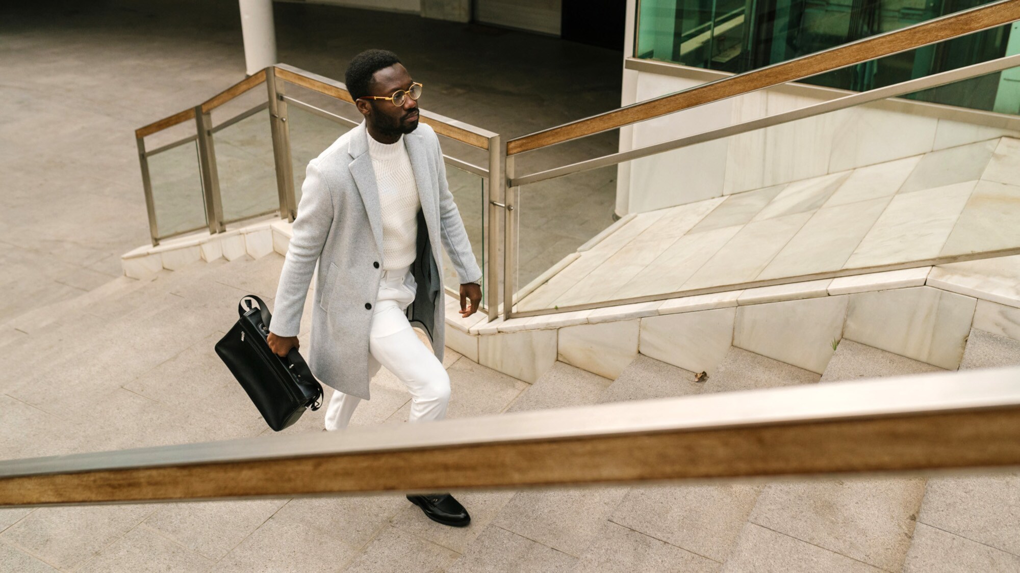 Ein elegant gekleideter Mann in einem hellen Outfit mit Wollmantel und schwarzer Aktentasche läuft eine Außentreppe hoch.
