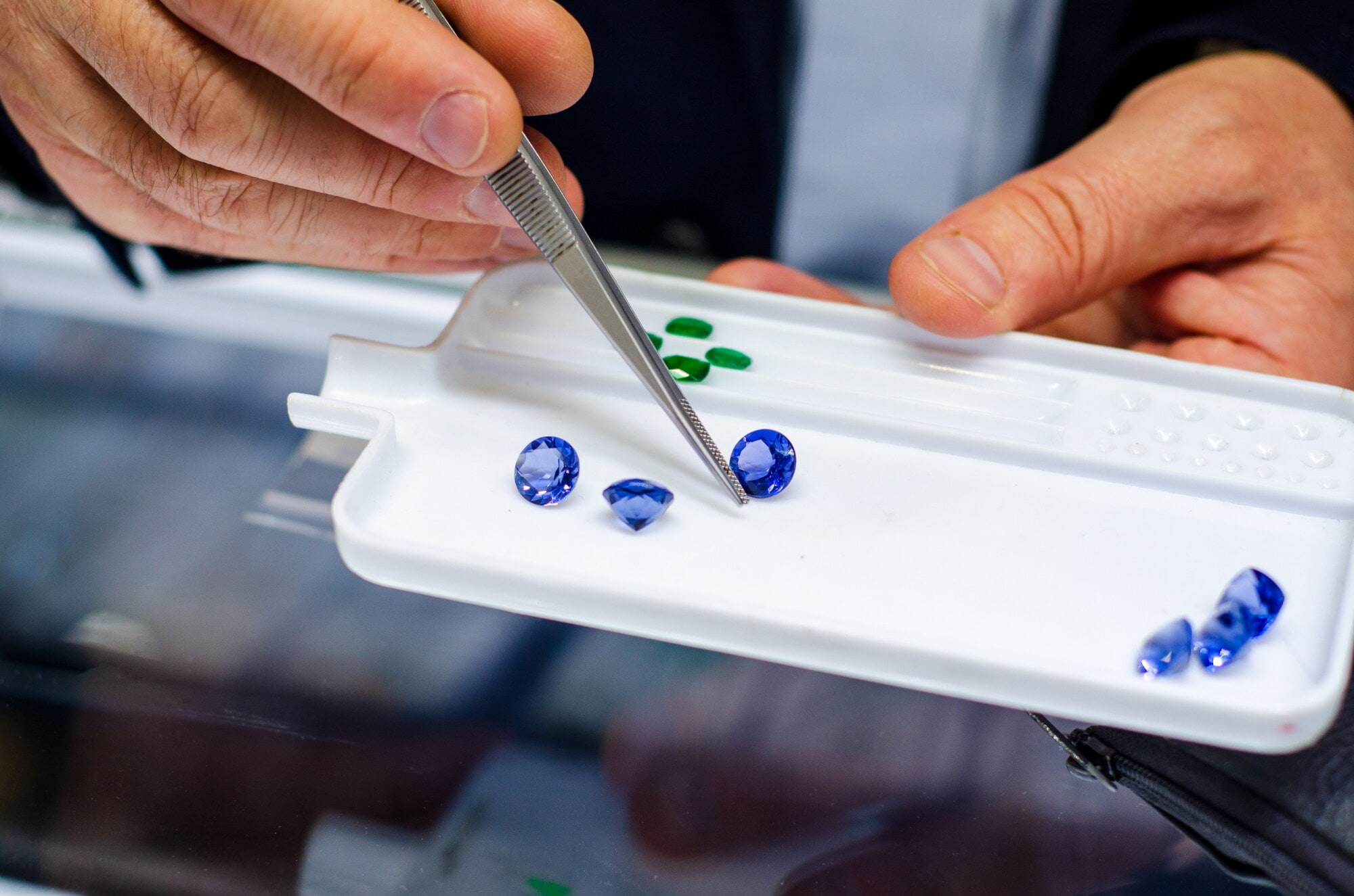 Mehrere blaue, geschliffene Edelsteine auf einem weißen Tablett, darüber eine Hand mit Pinzette. Mehrere blaue, geschliffene Edelsteine auf einem weißen Tablett, darüber eine Hand mit Pinzette.
