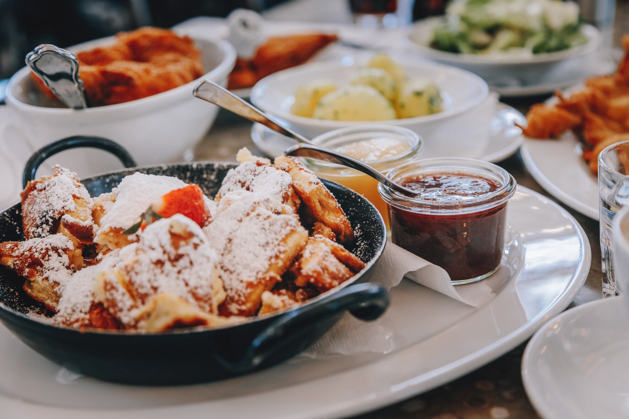 Ein schwarzer Topf mit Kaiserschmarrn auf einem weißen Teller, drumherum kleine Schälchen mit verschiedenen Leckereien.