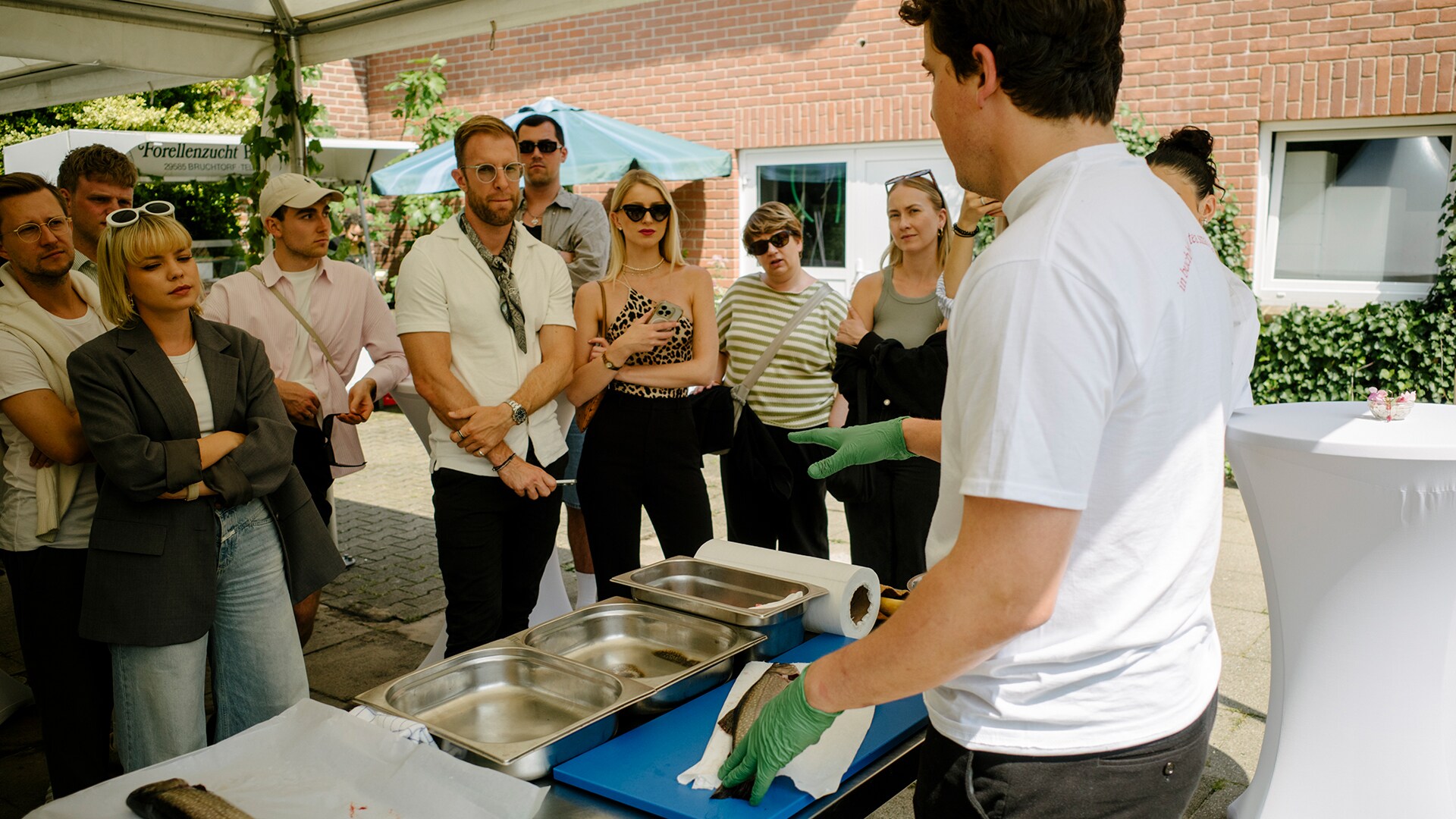 Eine Personengruppe beobachtet in einem Hof, wie ein Fisch von einem Koch filetiert wird. Eine Personengruppe beobachtet in einem Hof, wie ein Fisch von einem Koch filetiert wird.