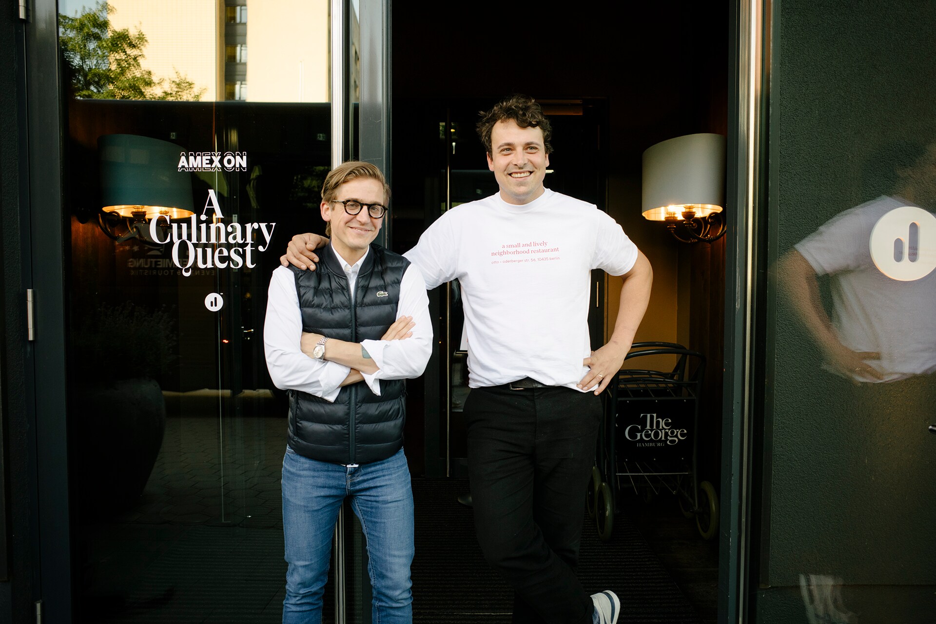 Zwei Männer posieren vor einer Glastür mit der Aufschrift „AMEX ON A Culinary Quest“ am Eingang eines Hotels.