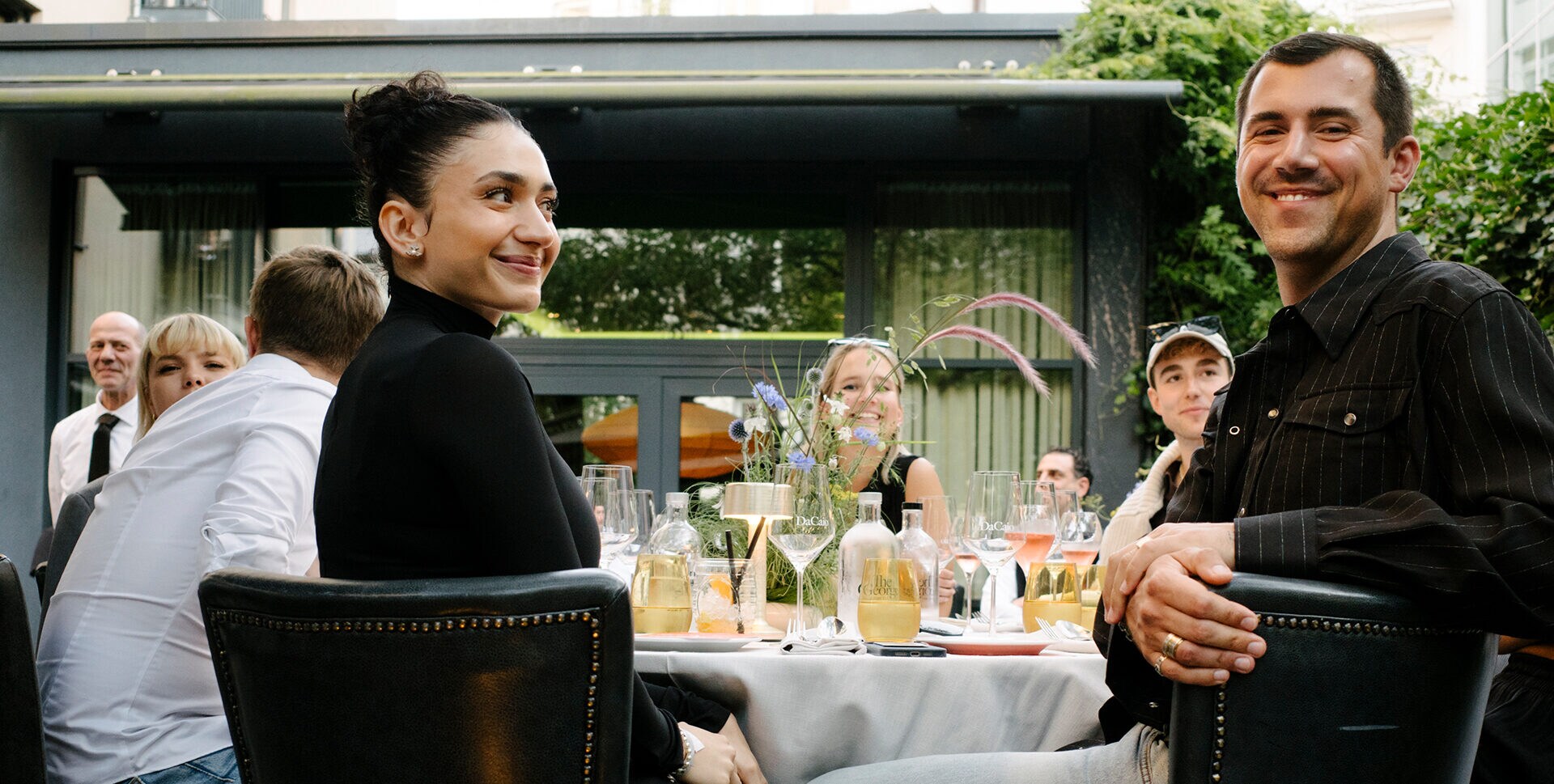 Mehrere Personen sitzen an einem Esstisch auf einer Hotelterrasse, zwei Personen im Vordergrundsind sind lächelnd in Richtung Kamera gewandt. Mehrere Personen sitzen an einem Esstisch auf einer Hotelterrasse, zwei Personen im Vordergrundsind sind lächelnd in Richtung Kamera gewandt.