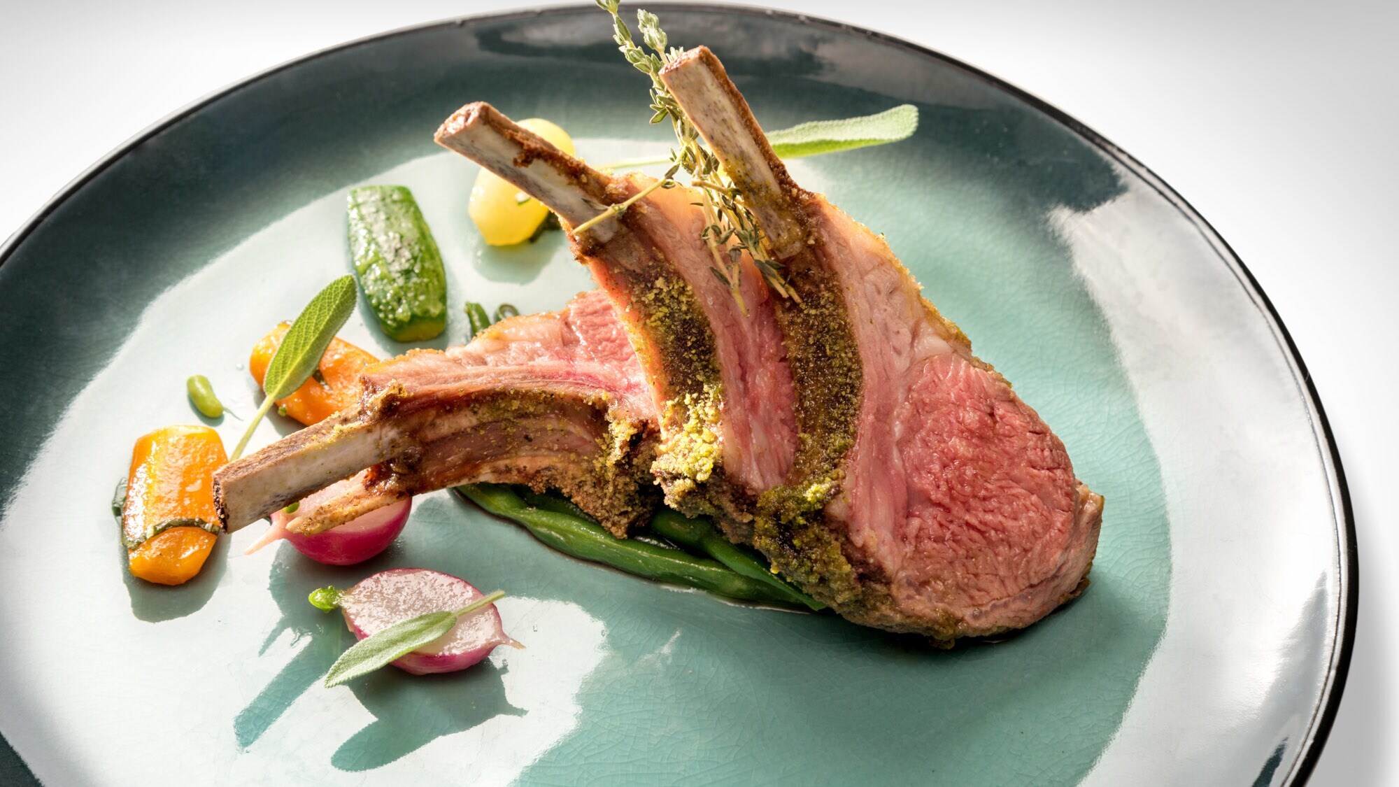 Lammkarree mit Kräuterkruste an Gemüse auf einem türkisfarbenen Teller.