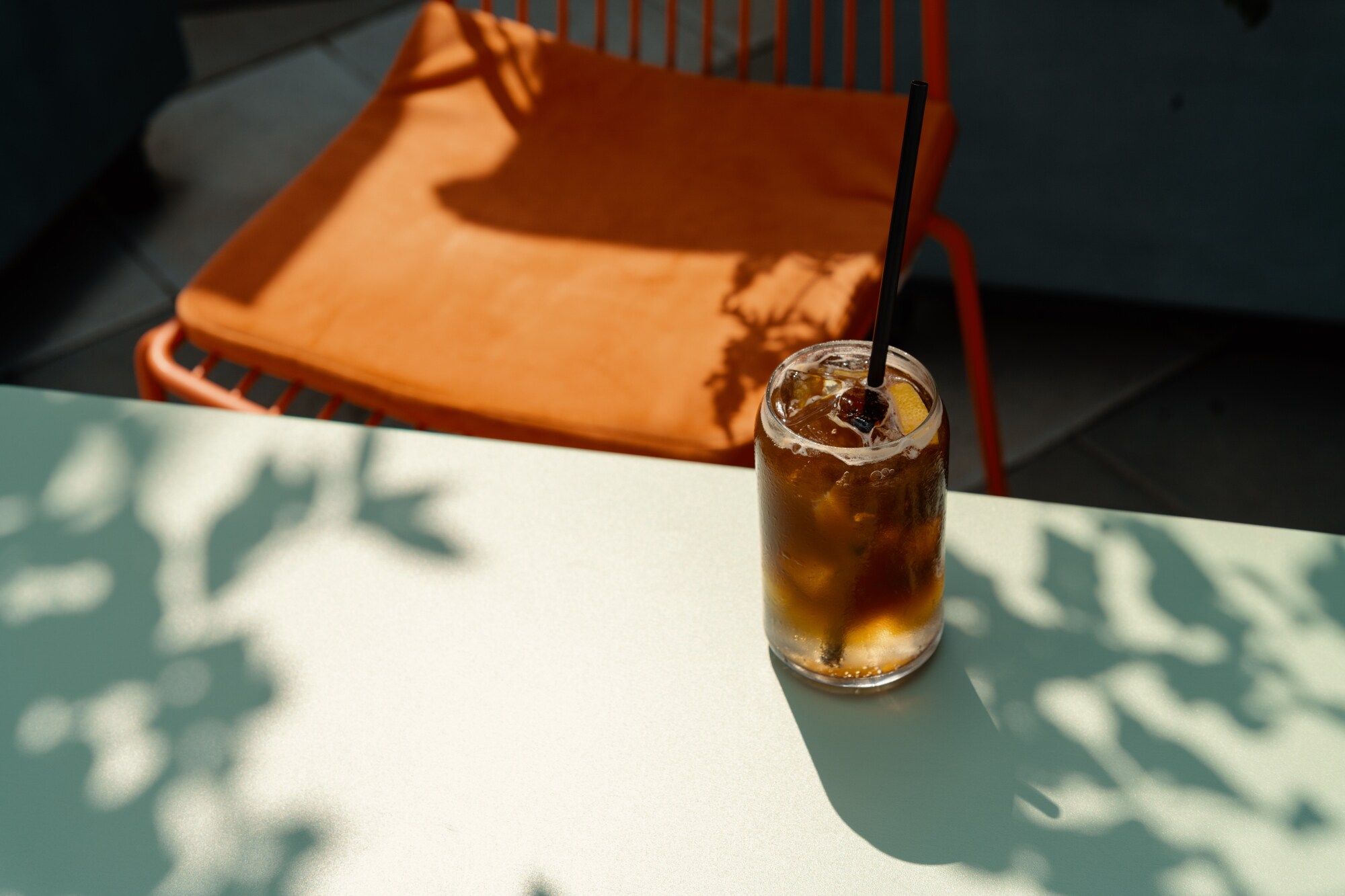 Ein Glas Espresso Tonic mit Strohhalm auf einem Tisch, dahinter steht ein orangefarbener Stuhl.