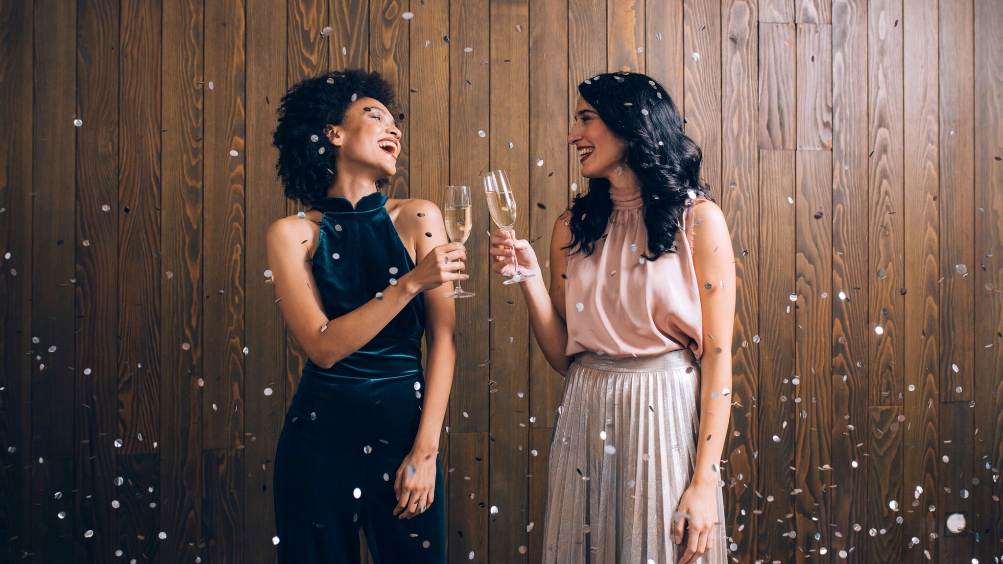 Zwei elegant gekleidete Frauen stehen im Konfettiregen vor einer Holzwand und stoßen mit Sektgläsern an. Zwei elegant gekleidete Frauen stehen im Konfettiregen vor einer Holzwand und stoßen mit Sektgläsern an.