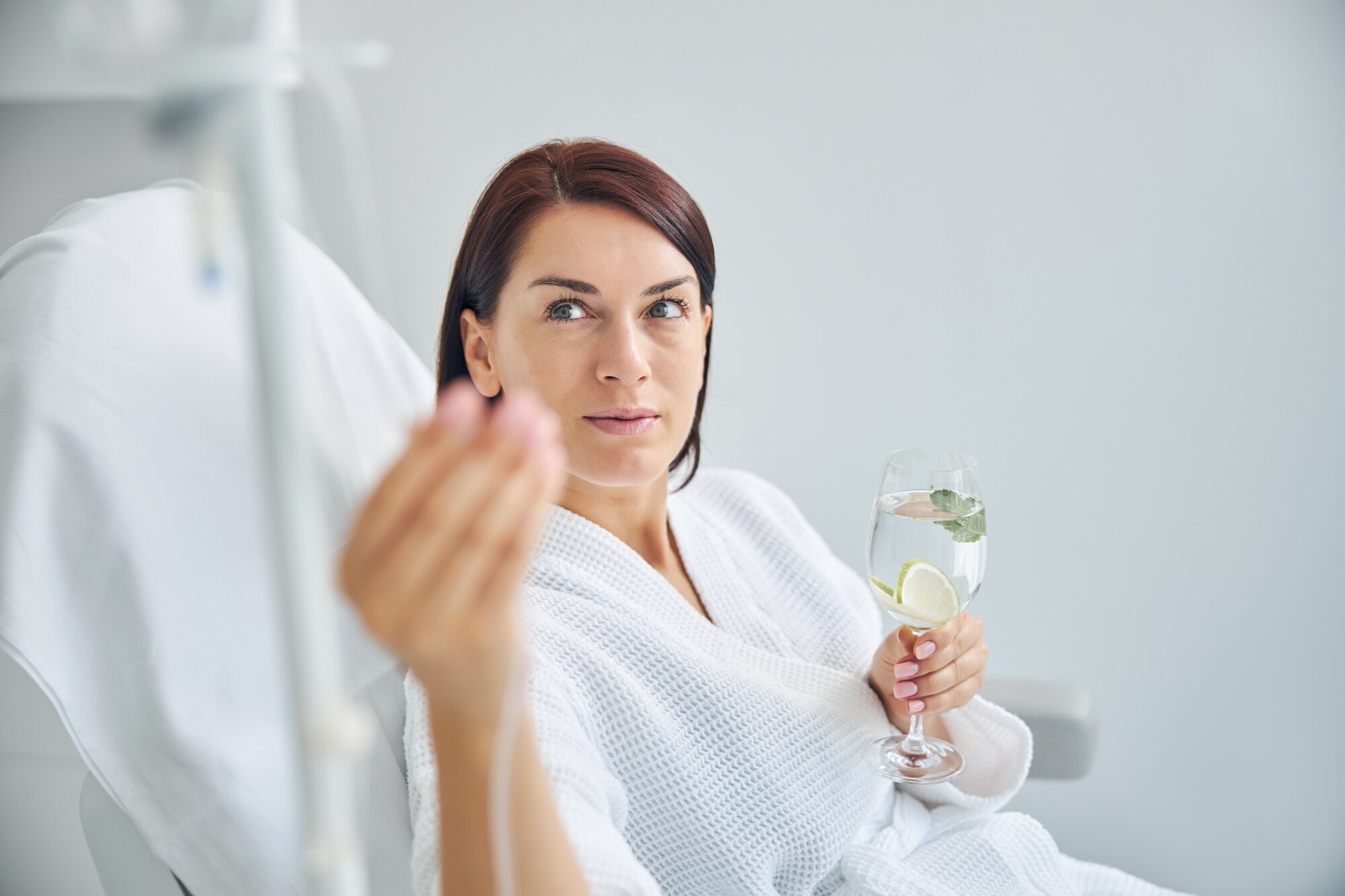 Frau in einer Schönheitsklinik reguliert ihre Vitamininfusion und hält ein Glas Wasser in der Hand.