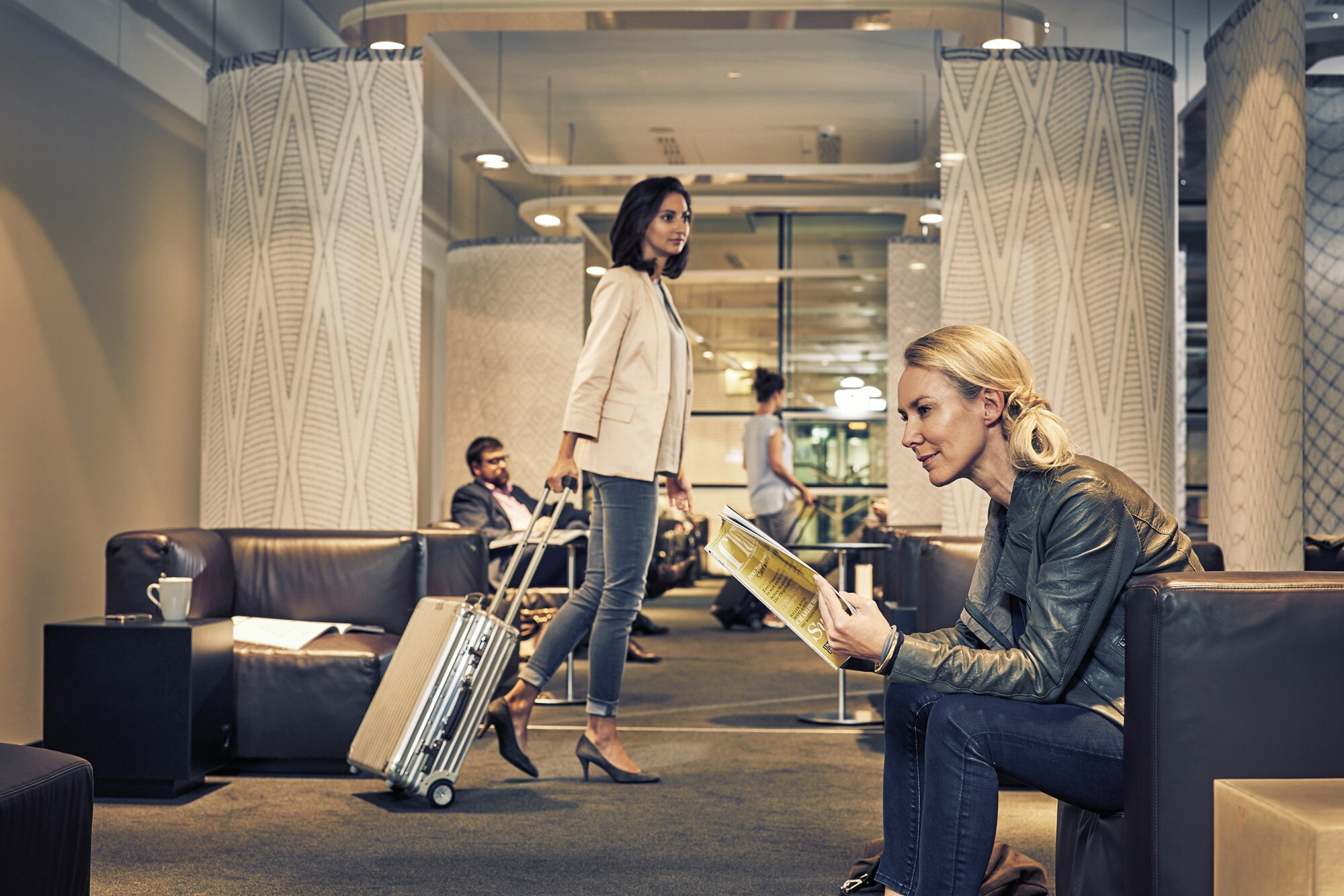 Eine Frau sitzt in einem Ledersessel und blättert in einem Magazin, im Hintergrund sind weitere Menschen zu sehen