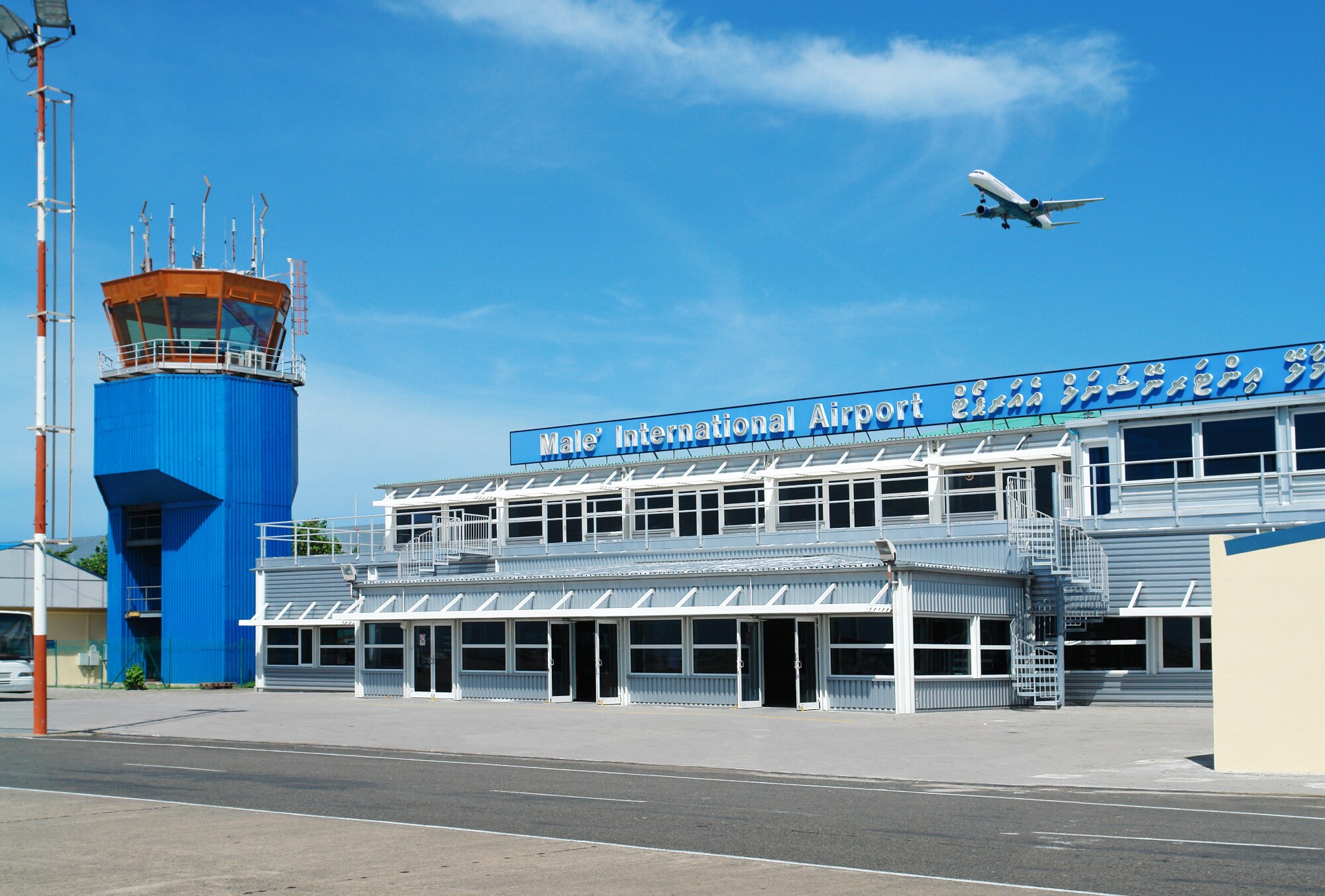 Ein Flugzeug fliegt am blauen Himmel über einen kleines Flughafengebäude mit blauem Kontrollturm.