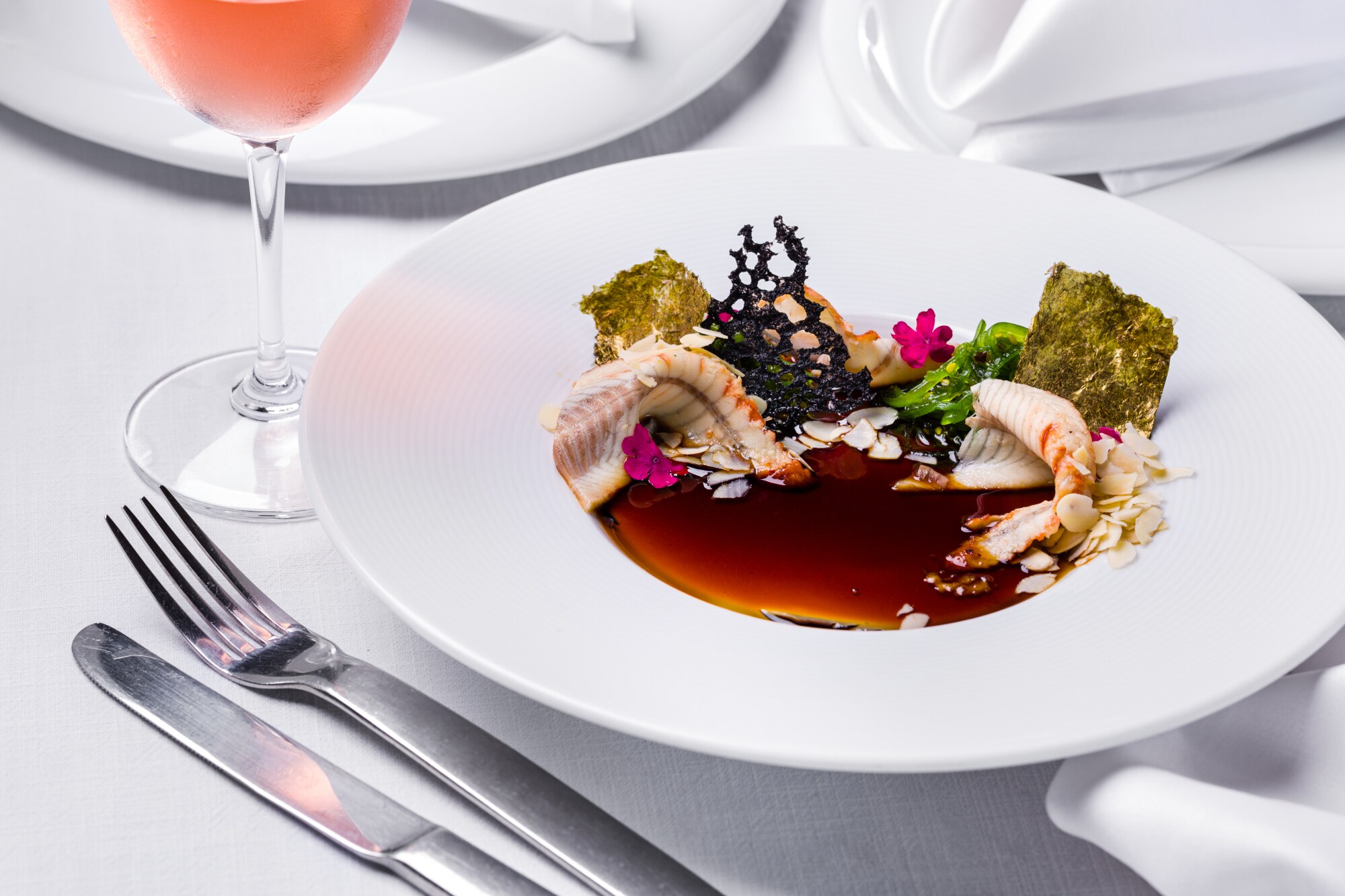 Ein kreativ angerichtetes Fischgericht auf einem weißen Teller auf einem weiß eingedeckten Tisch.
