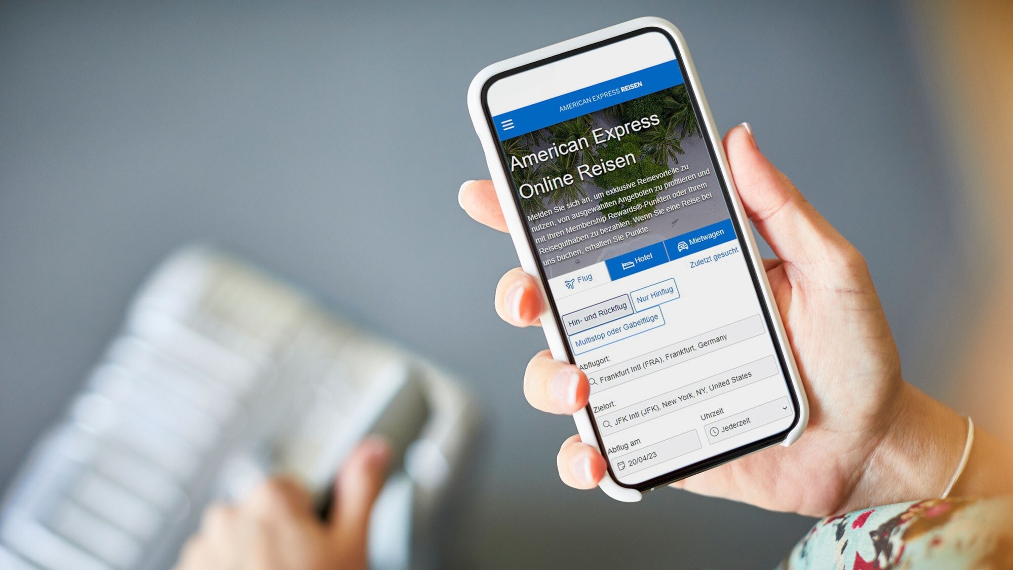 Ein Smartphone mit dem American Express Online-Reiseportal auf dem Display wird von einer Hand über einen Koffer gehalten.