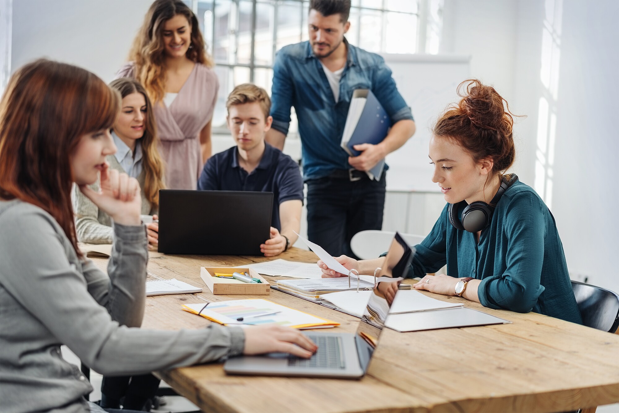 Co-Working: Der Booster für deine Work-Life-Balance Mehrere Personen befinden sich in einem hellen loftartigen Coworking-Space und arbeiten an einem Tisch zusammen am Laptop und mit Unterlagen.