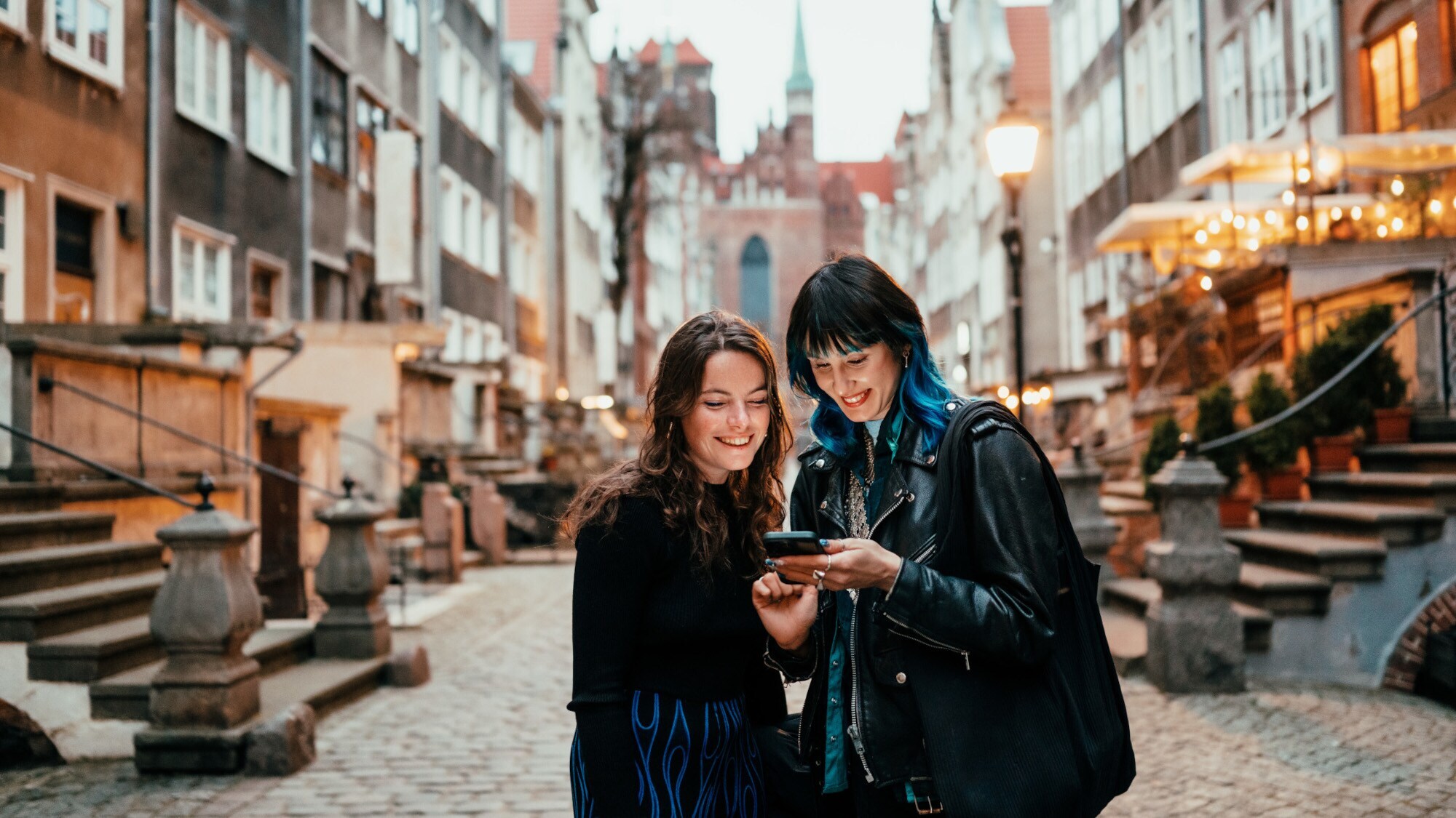 Zwei junge Frauen stehen in einer kopfsteingepflasterten Straße und blicken auf ein Smartphone, das eine von ihnen in der Hand hält.