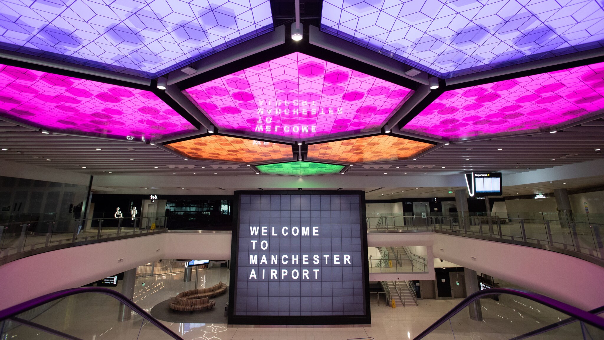 Menschenleere Flughafenhalle mit einem Bildschirm im Mittelpunkt, auf dem „Welcome to Manchester Airport“ steht und bunt erleuchteten LED-panels an der Decke. Menschenleere Flughafenhalle mit einem Bildschirm im Mittelpunkt, auf dem „Welcome to Manchester Airport“ steht und bunt erleuchteten LED-panels an der Decke.
