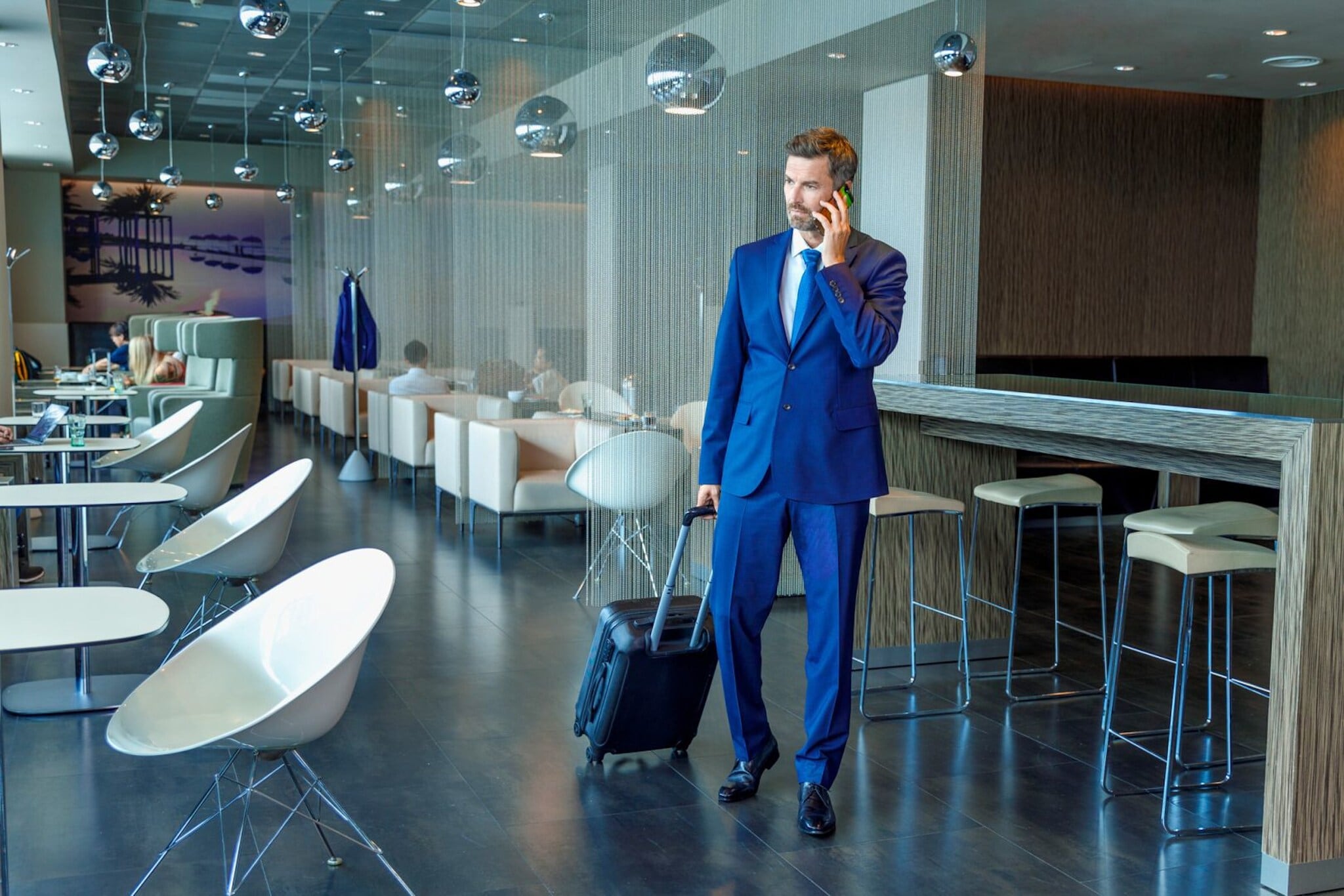 Ein Geschäftsmann im blauen Anzug und mit Trolley telefoniert stehend in einer modernen Flughafen-Lounge. Ein Geschäftsmann im blauen Anzug und mit Trolley telefoniert stehend in einer modernen Flughafen-Lounge.
