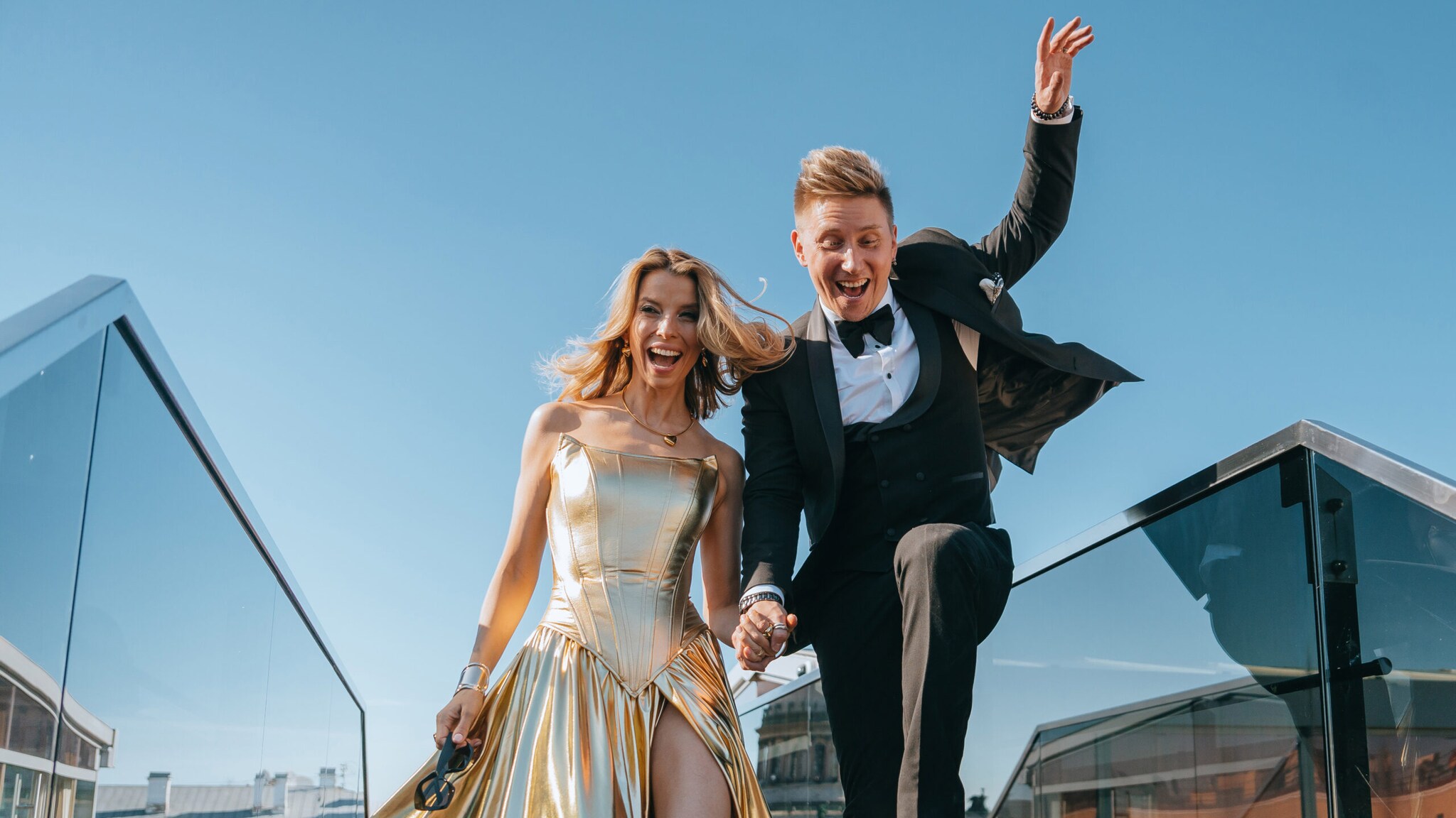 Ein lachendes Paar in Abendgarderobe läuft händchenhaltend in einem urbanen Umfeld.