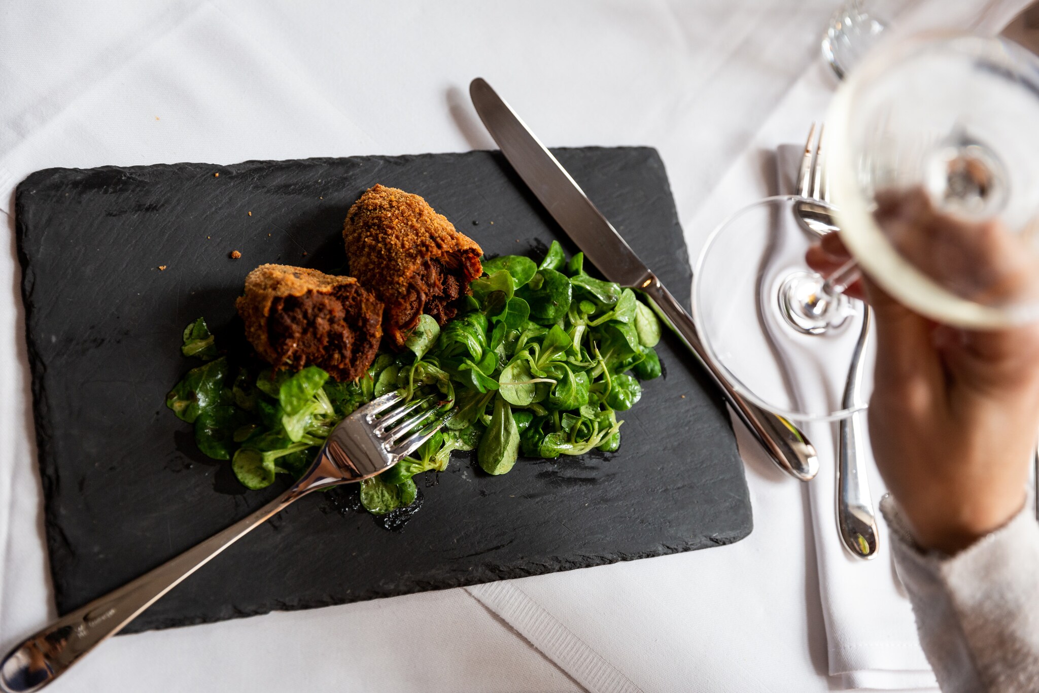 Aufsicht eines panierten Fleischgerichtes an Feldsalat auf einer Schieferplatte auf einem edel gedeckten Tisch, im Vordergrund eine Hand mit einem Glas Weißwein.