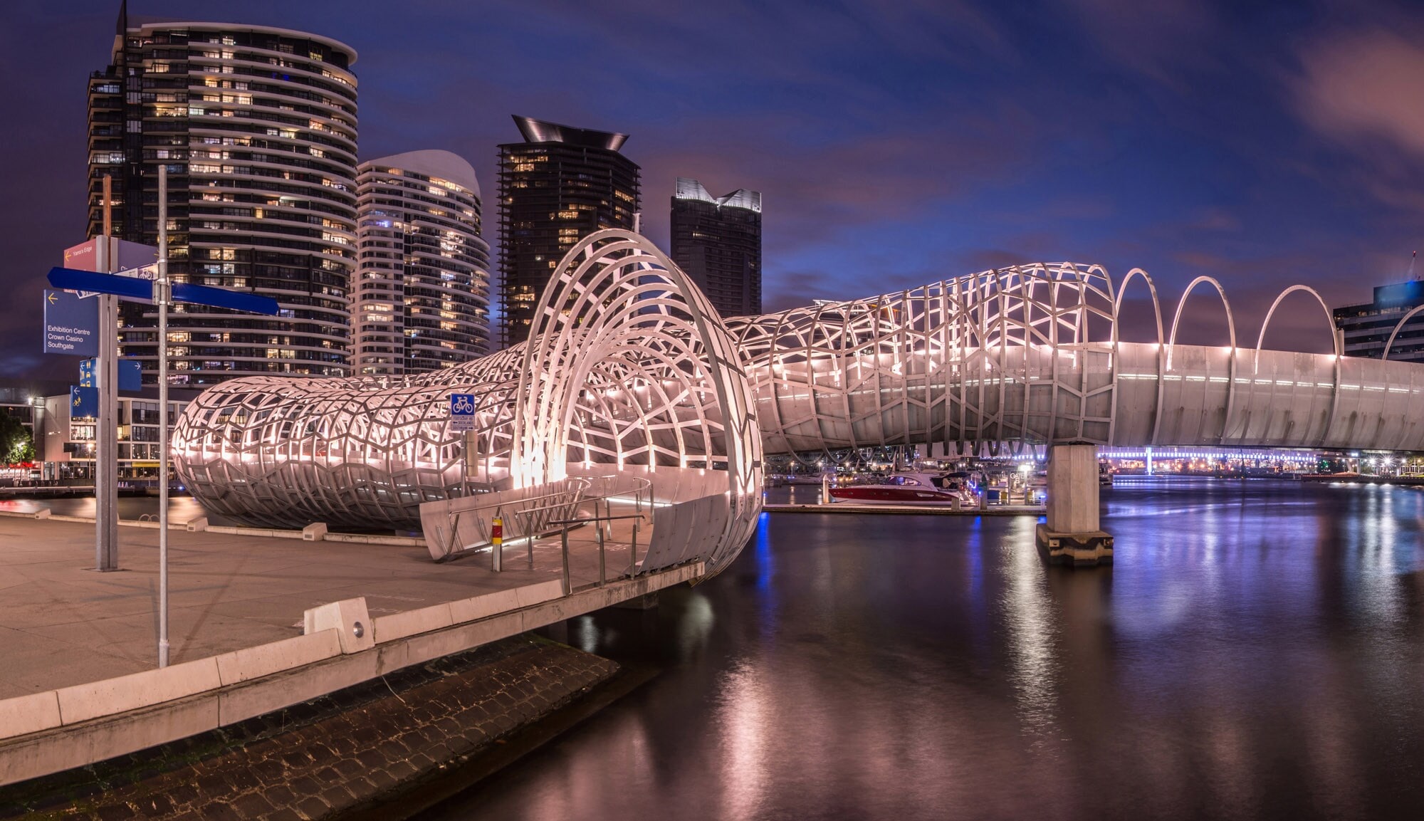 Geschwungene Brücke aus einer kunstvollen Stahlkonstruktion an der Promenade eines modernen Hafenviertels bei Nacht. Geschwungene Brücke aus einer kunstvollen Stahlkonstruktion an der Promenade eines modernen Hafenviertels bei Nacht.