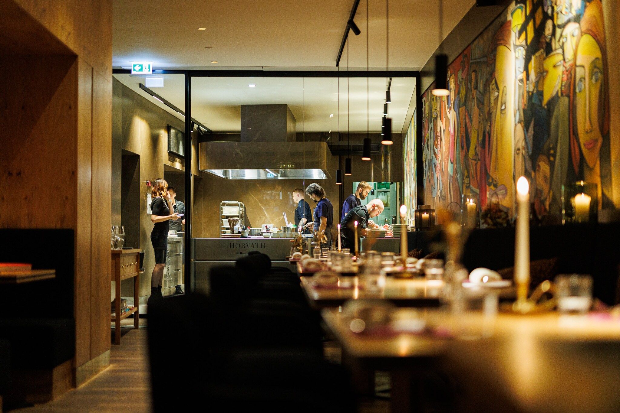 Personen in einer offenen Restaurantküche, davor Speiseraum mit eingedeckten Tischen bei Kerzenschein.