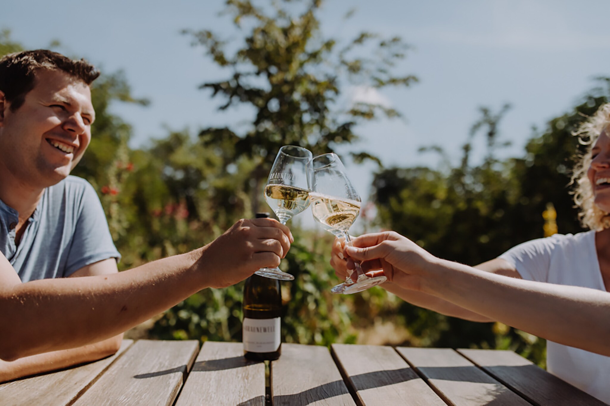 Zwei Personen beim Anstoßen mit Weißweingläsern an einem Holztisch im Freien bei Sonnenschein.