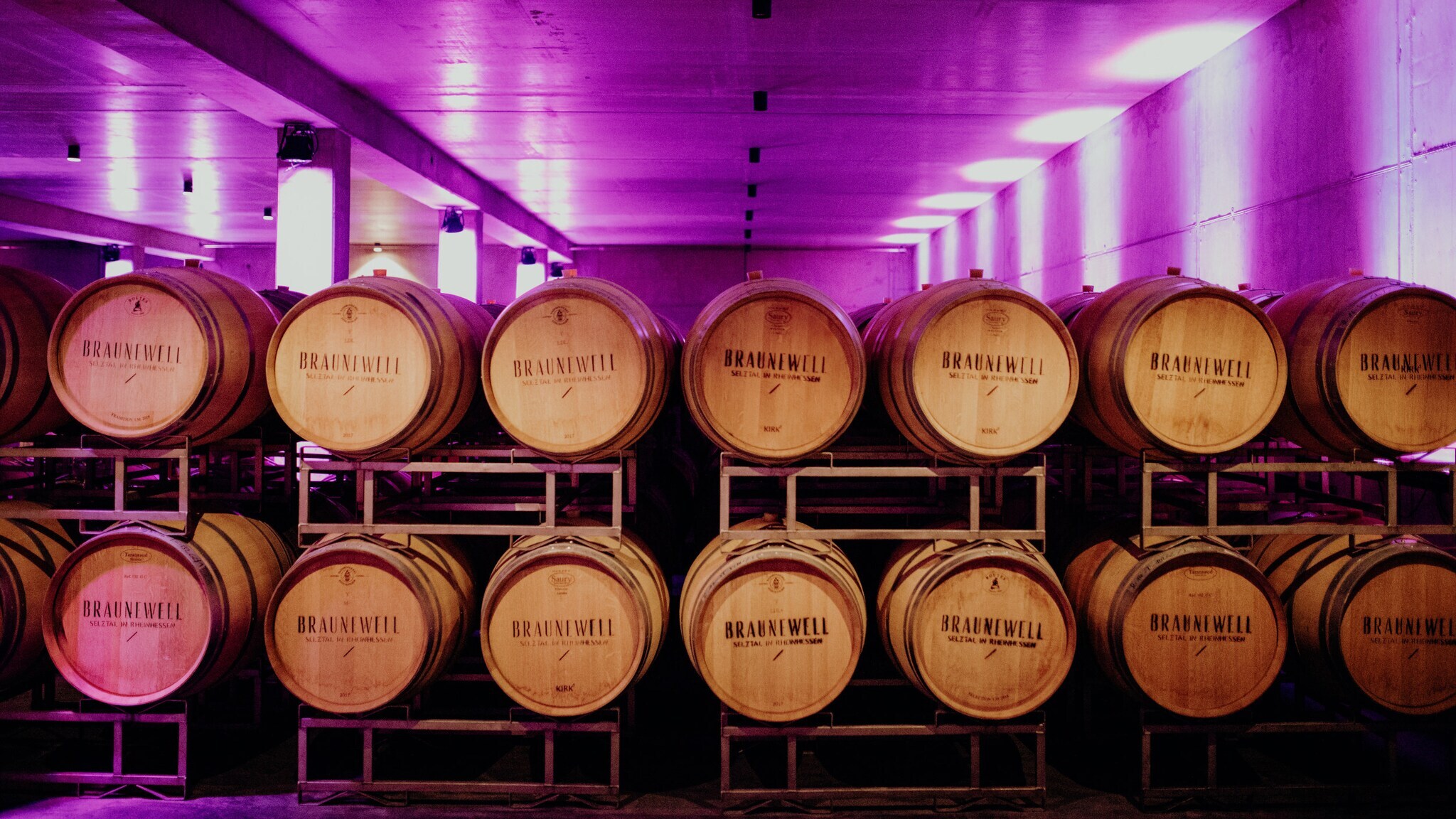 Gestapelte Barriquefässern in einem modernen Weinkeller mit pink beleuchteten Betonwänden.