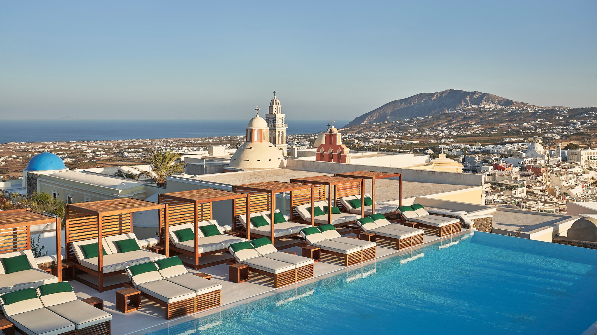 Dachterrasse mit zahlreichen Sonnenliegen am Infinity-Pool auf Santorini, im Hintergrund Meeresküste. Dachterrasse mit zahlreichen Sonnenliegen am Infinity-Pool auf Santorini, im Hintergrund Meeresküste.