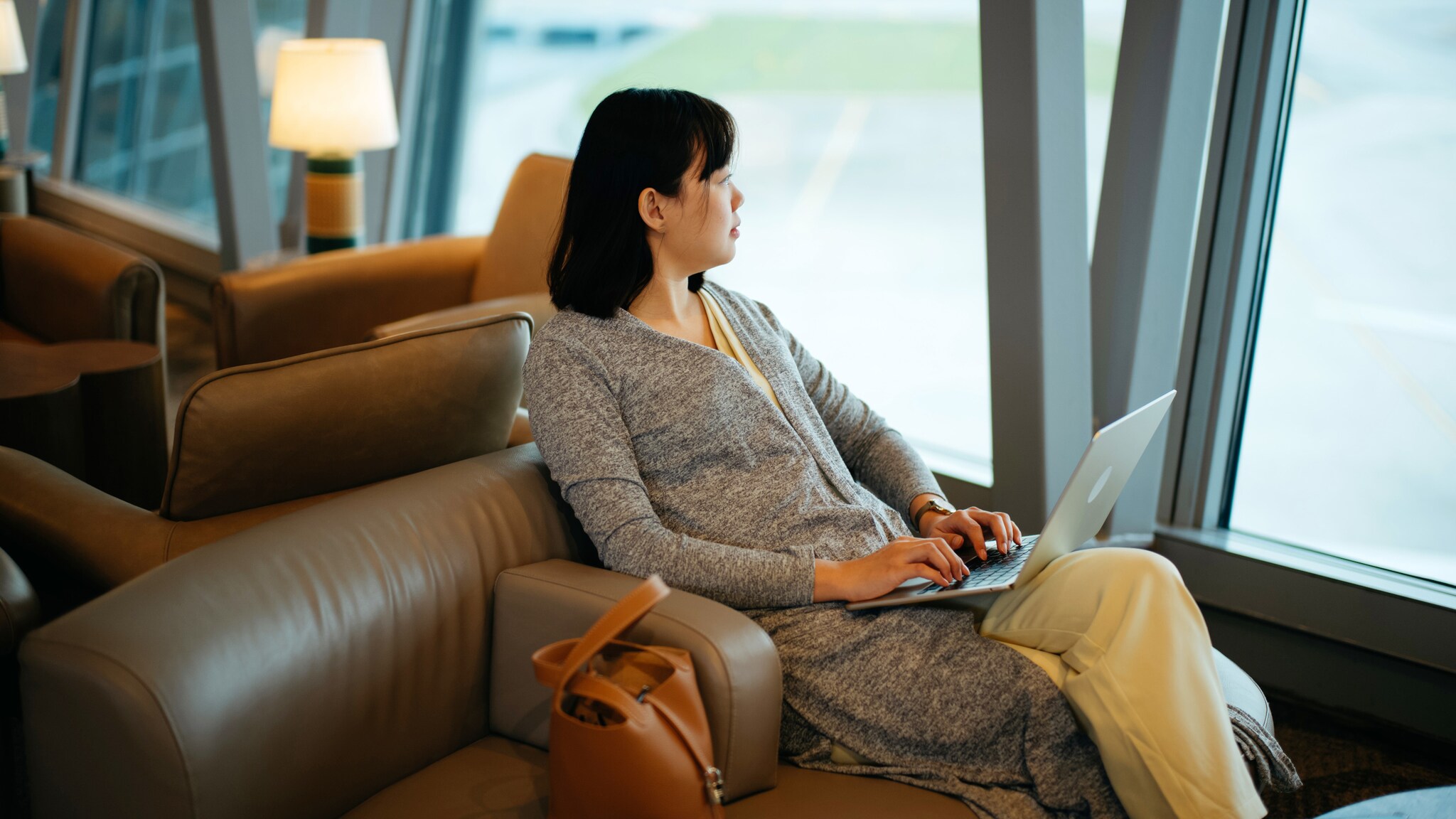 Eine Frau sitzt mit Laptop in einem Ledersessel am Fenster einer Flughafen-Lounge und schaut aufs Rollfeld.